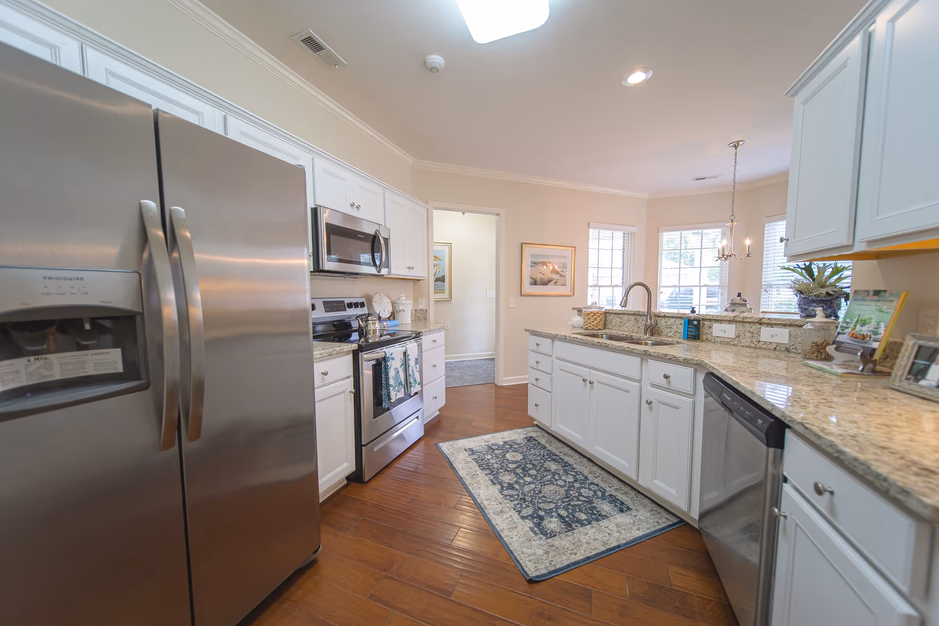 A modern kitchen with stainless steel appliances including a refrigerator, microwave, stove, and dishwasher. The kitchen features white cabinets, granite countertops, a sink with a faucet, a decorative rug on the wooden floor, and windows letting in natural light.