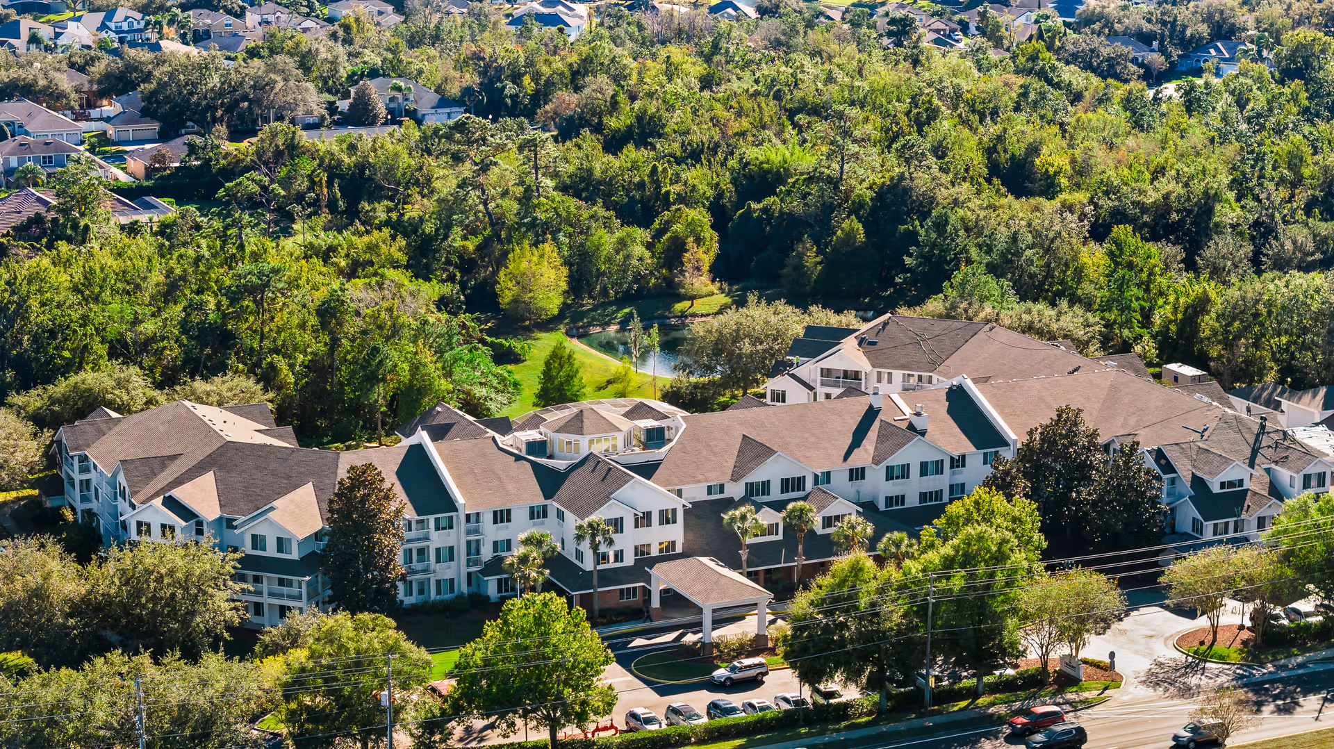 Aerial view of The Tremont - A Provincial Senior Living Community, showing a large multi-wing building surrounded by trees and greenery with a parking area and road in the foreground.