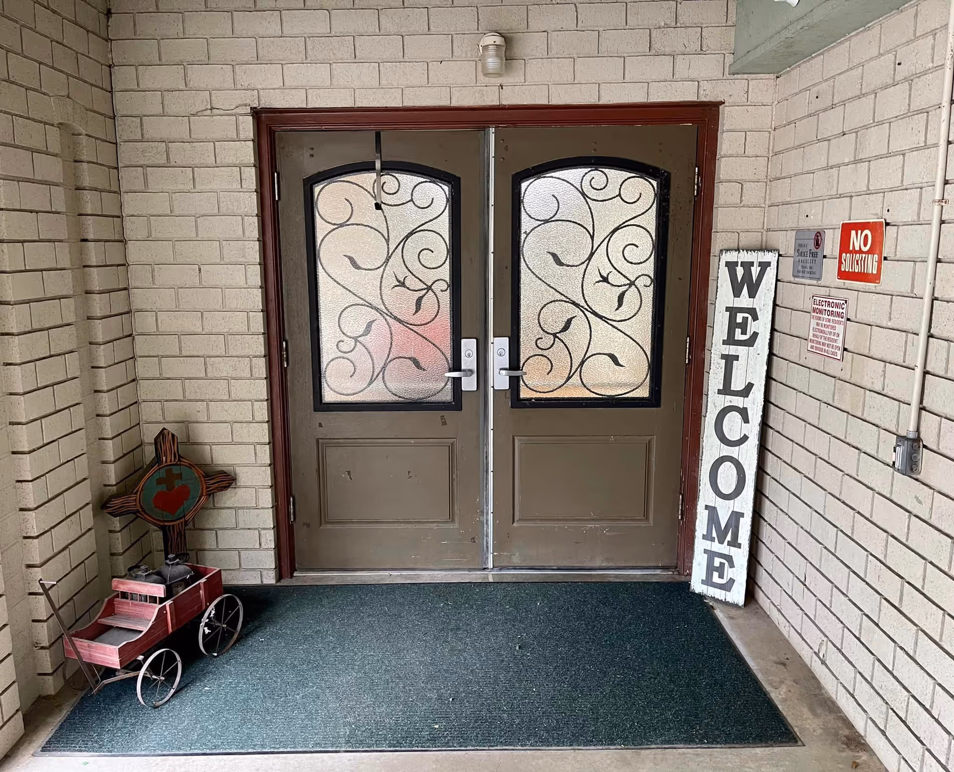 Double entrance doors with decorative frosted glass, a leaning 'WELCOME' sign, a small red wagon, and a 'No Soliciting' notice by the doorway.
