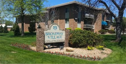Exterior view of Broadway Village, a brick senior living facility building with multiple windows and a blue awning. In front of the building is a landscaped area with green grass, bushes, trees, and a stone sign that reads 'Broadway Village A Total Living Experience.'