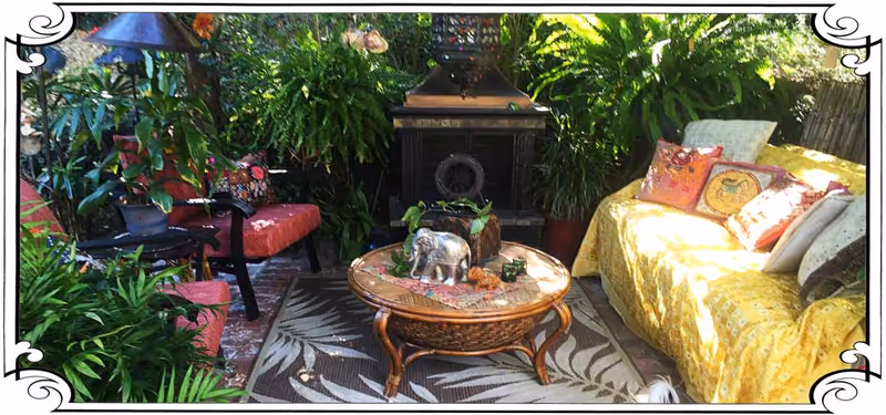 A cozy outdoor seating area with lush green plants surrounding a red cushioned bench on the left and a yellow couch with multiple colorful pillows on the right. In the center, there is a round wooden coffee table with decorative items including a small elephant figurine. A black fireplace is visible in the background.