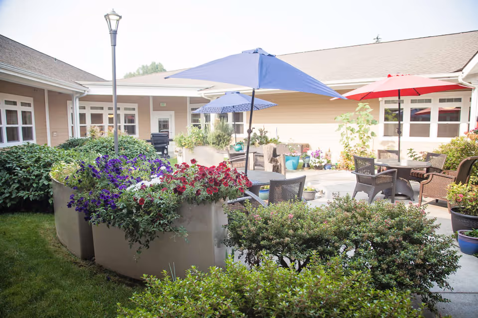 Outdoor courtyard area of a senior living facility with large planters filled with colorful flowers, patio tables and chairs with blue and red umbrellas, surrounded by beige building walls and windows.