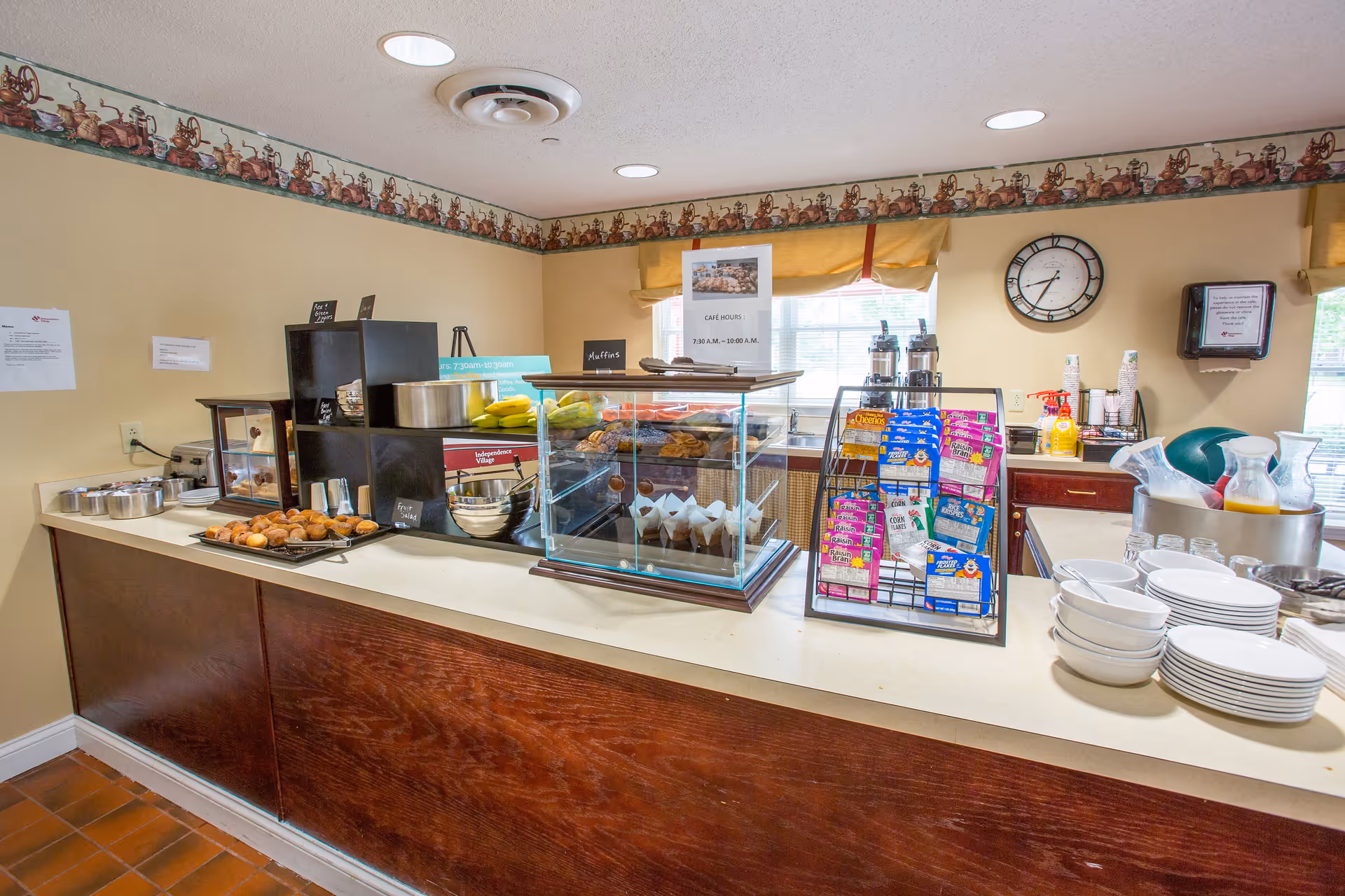 A breakfast or snack counter in a senior living facility with muffins, bananas, cereal boxes, plates, bowls, and coffee dispensers. The room has beige walls with a decorative border near the ceiling, a clock on the wall, and a window with a yellow valance.