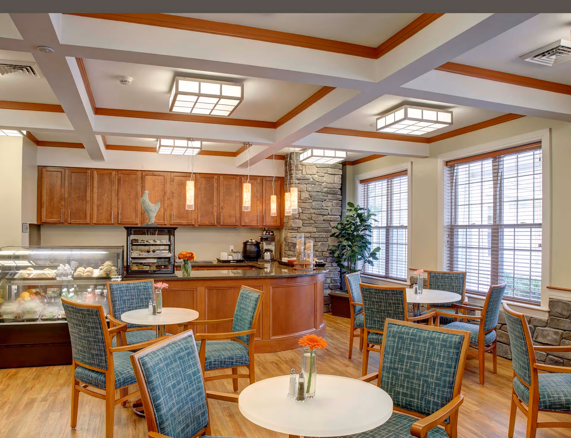 A bright dining area with round white tables and blue cushioned chairs. The room features wooden flooring, large windows with blinds, and a counter with wooden cabinets and pendant lights. There is a display case with food items and a stone accent wall with a potted plant nearby.