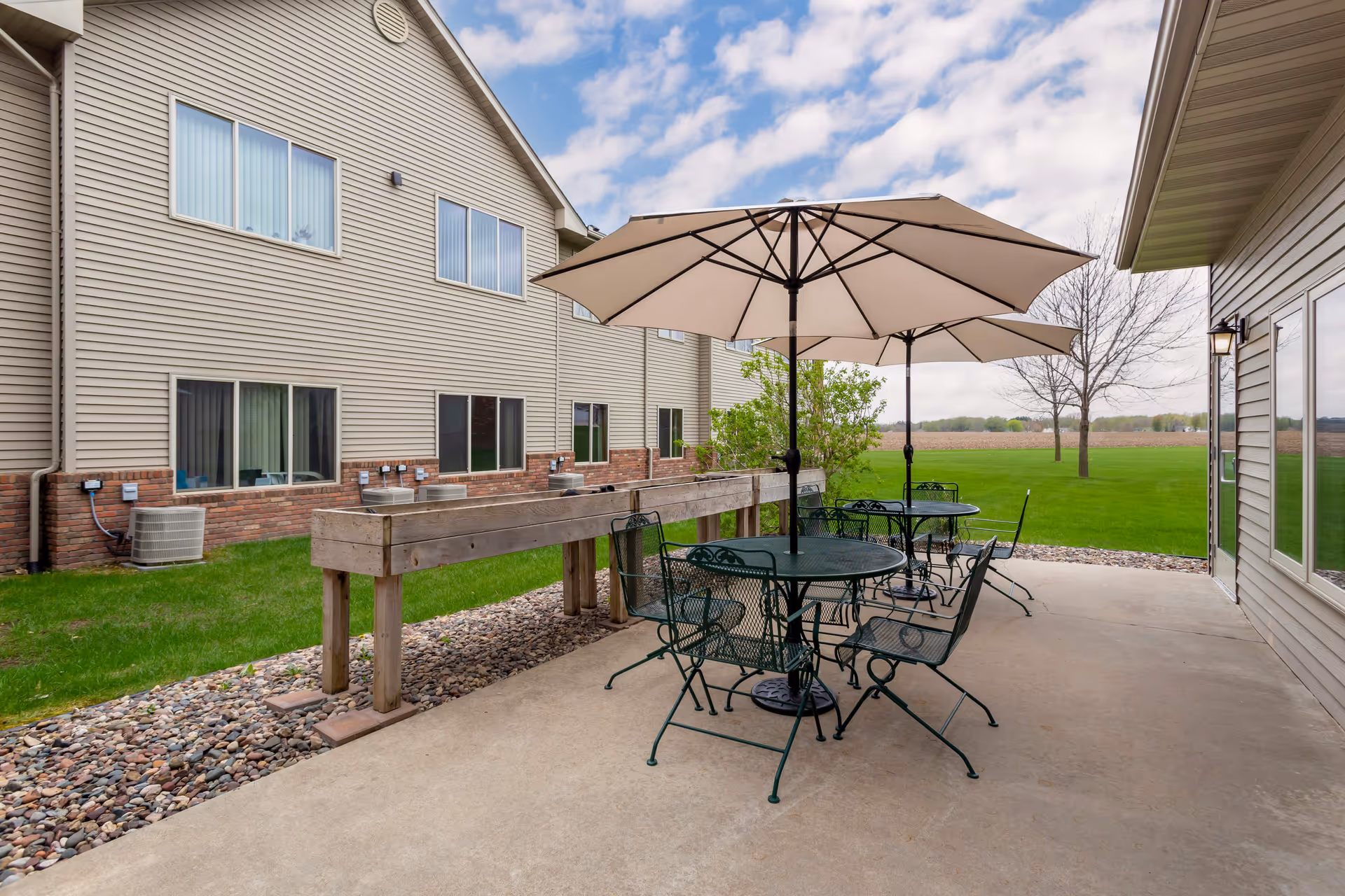 Outdoor patio area at South Grove Lodge Senior Living with metal tables and chairs under large beige umbrellas, adjacent to a beige building with multiple windows. The patio overlooks a green lawn and open field under a partly cloudy sky.