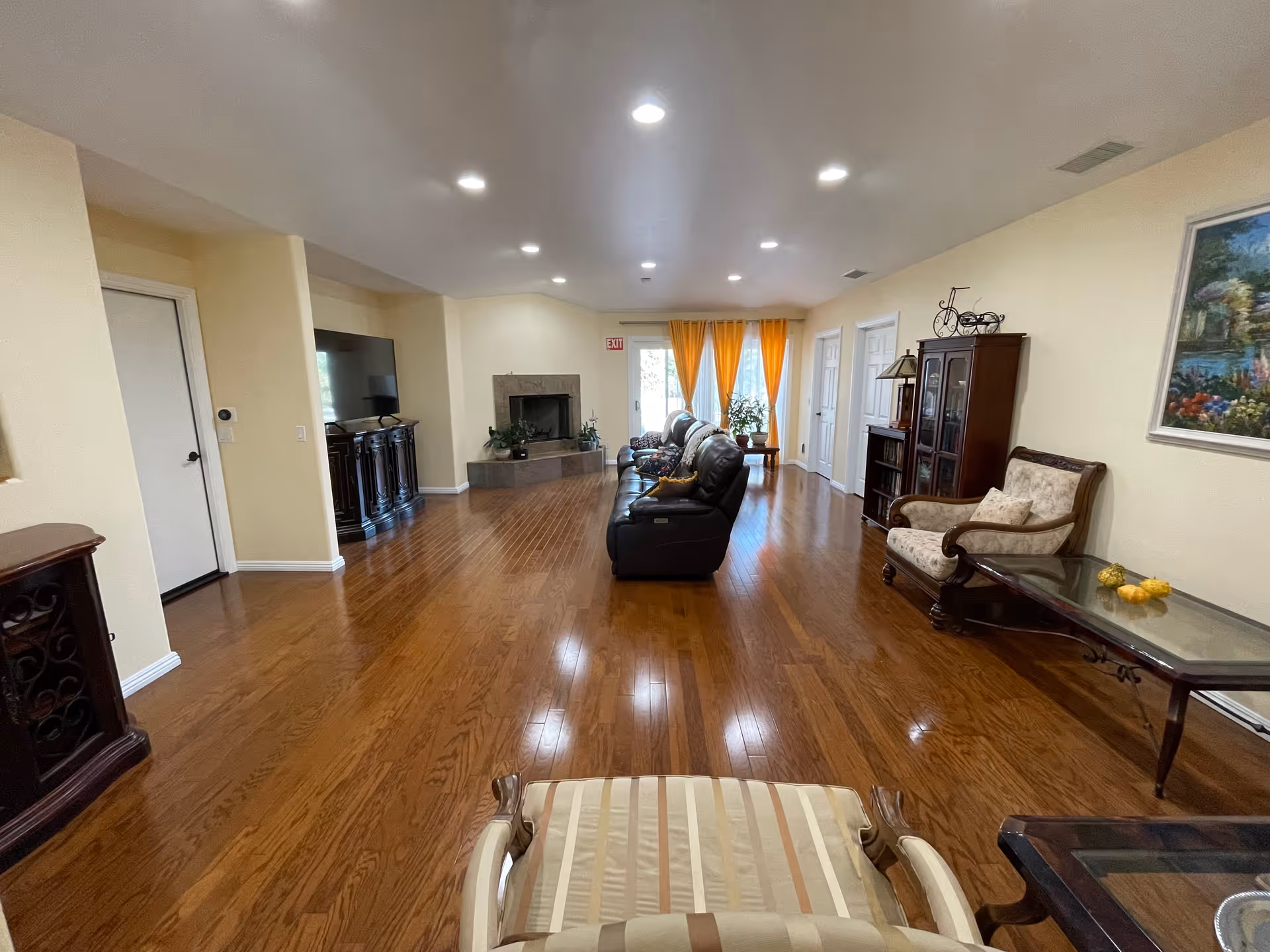 A spacious living room with polished wooden floors, beige walls, and recessed ceiling lights. The room features a row of dark leather sofas facing a fireplace and a large flat-screen TV on a wooden cabinet. There are large windows with yellow curtains at the far end, letting in natural light. On the right side, there is a wooden cabinet with glass doors, an upholstered armchair, and a glass-top coffee table. A painting hangs on the wall above the armchair.