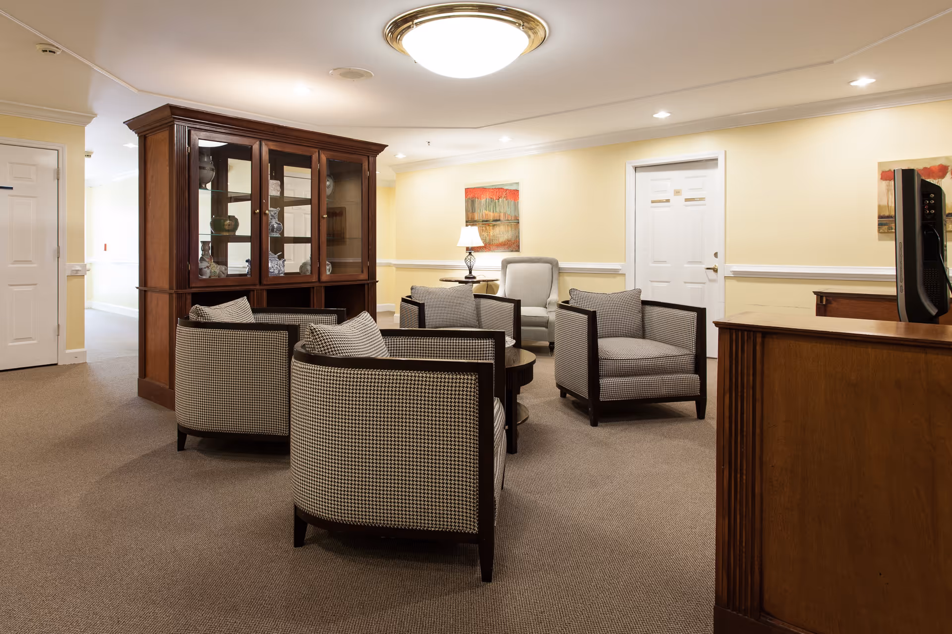 A cozy senior living common area with four cushioned armchairs arranged around a small round table. There is a wooden display cabinet with glass doors containing decorative items, a single armchair with a side table and lamp, and a TV on a wooden stand. The walls are painted light yellow with white trim, and there are two paintings hanging on the walls.
