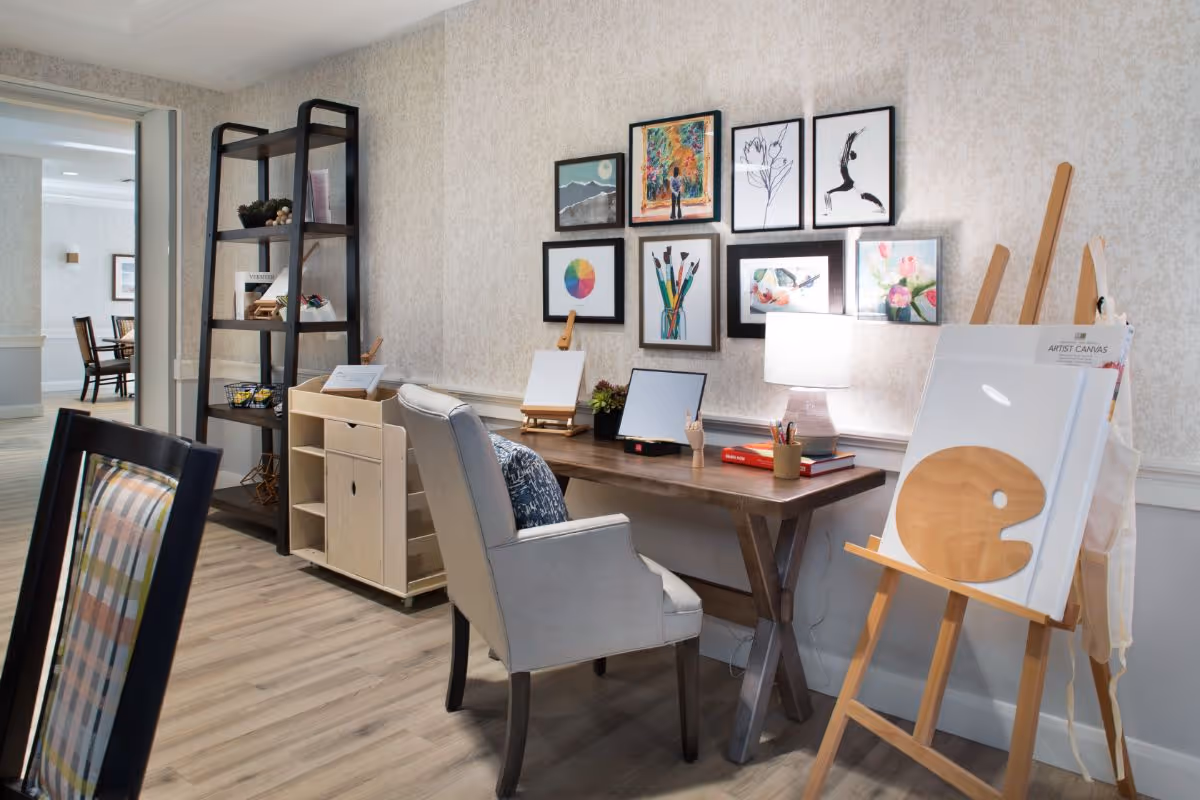 A cozy art corner in a senior living facility with a wooden table, a comfortable upholstered chair, and an easel holding a blank artist canvas. The wall behind the table is decorated with framed colorful artwork and sketches. A tall black shelving unit and a small wooden storage cart are positioned nearby. The room has light-colored walls and wood flooring, with a glimpse of a dining area visible through an open doorway.