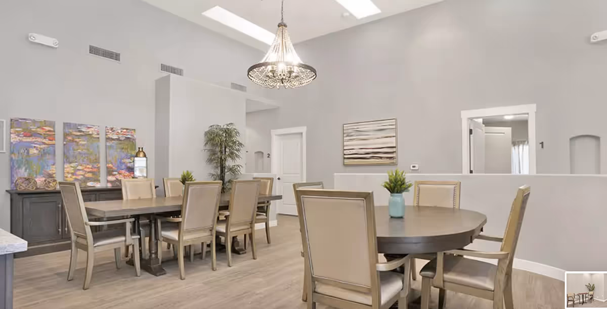 A spacious dining room with two wooden dining tables surrounded by beige cushioned chairs. The room features light gray walls, a modern chandelier hanging from a high ceiling, colorful artwork on the walls, and a potted plant near the corner. There are two white doors labeled with numbers 1 and 2.