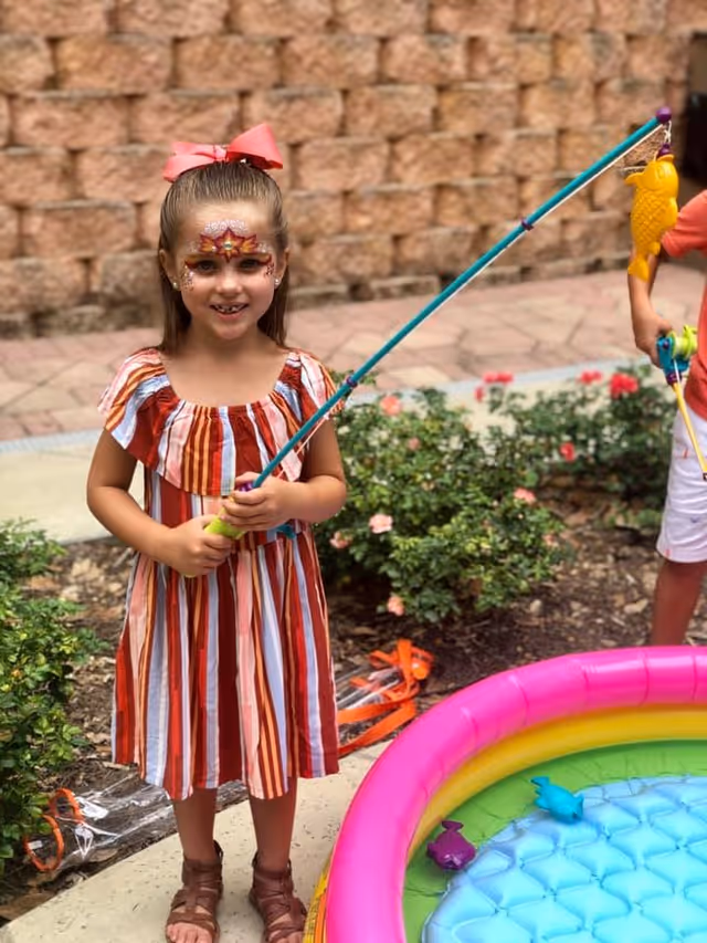 A young girl with face paint and a pink bow in her hair is holding a fishing pole with a yellow fish attached. She is standing next to a small inflatable pool with colorful toy fish inside. There is greenery and a stone wall in the background.