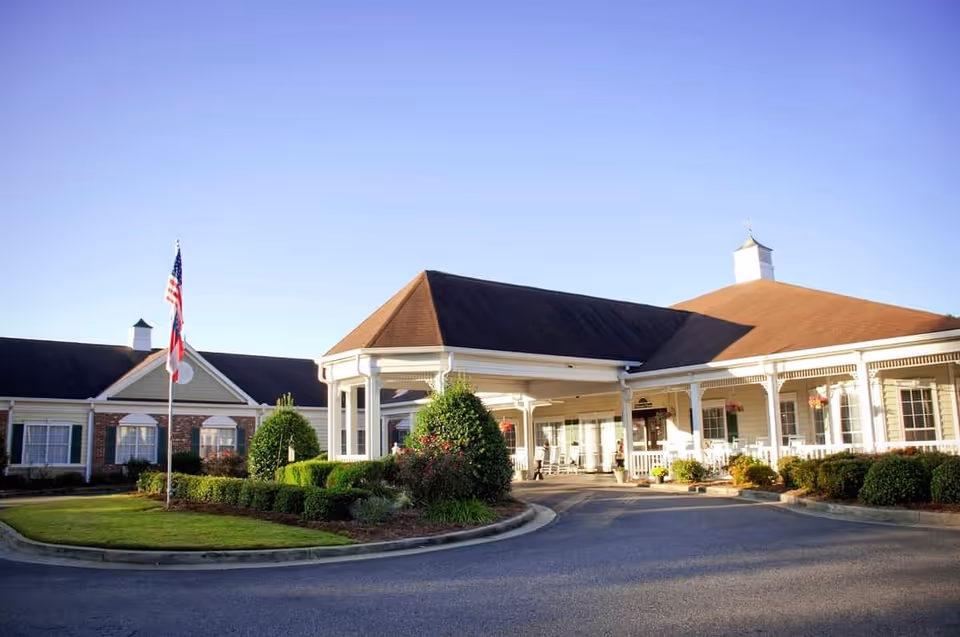 Front entrance of a single-story senior living building with a covered porte-cochère, American flag, and landscaped circular driveway.