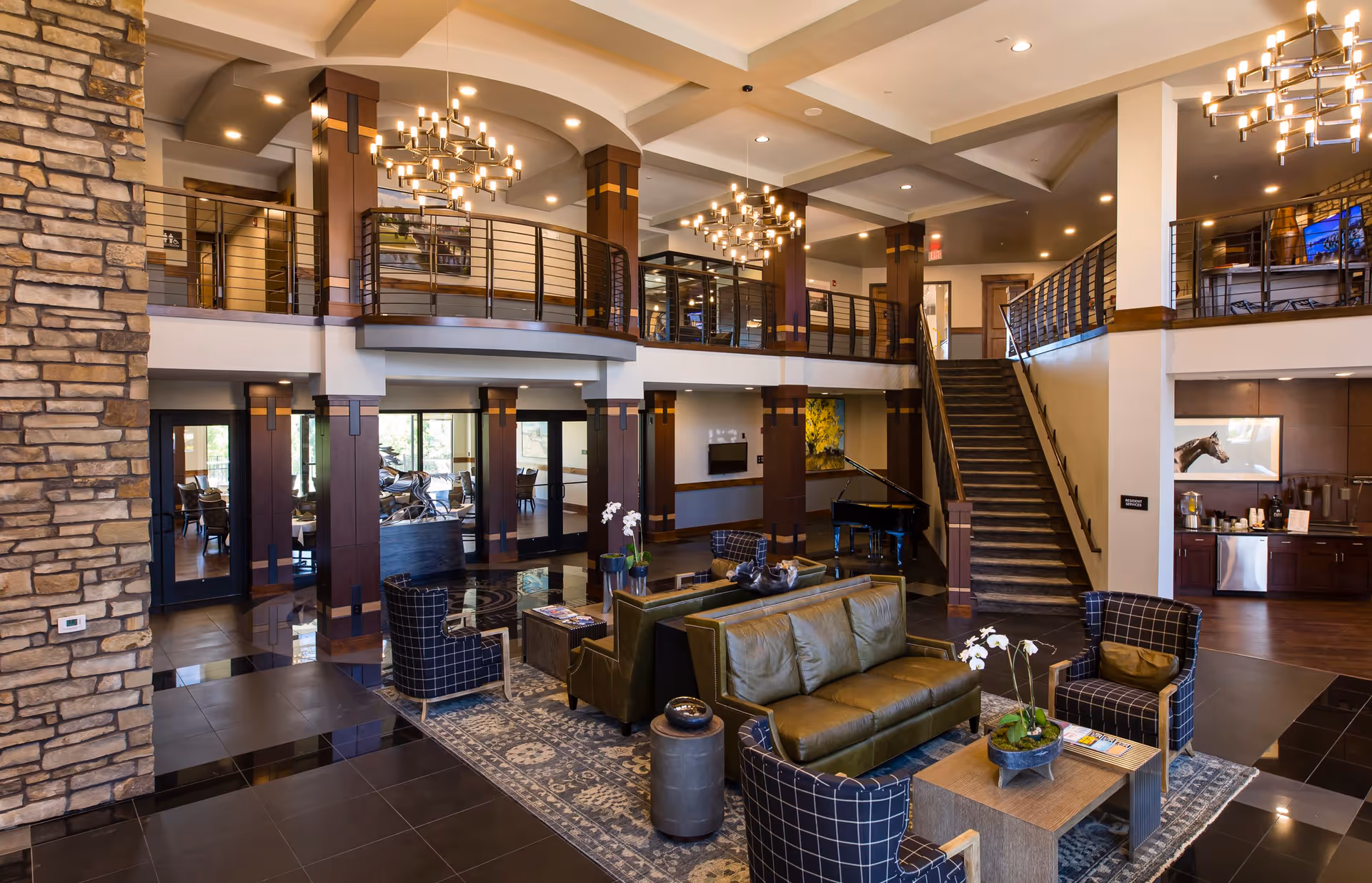Spacious and elegant common area in Fairways of Ironhorse Independent Living featuring a seating area with green leather sofas and patterned armchairs around a wooden coffee table with plants. The room has a high ceiling with modern chandeliers, a stone accent wall, a grand piano, and a staircase leading to an upper balcony with railings. There is also a kitchenette area with a coffee station and a framed horse picture on the wall.