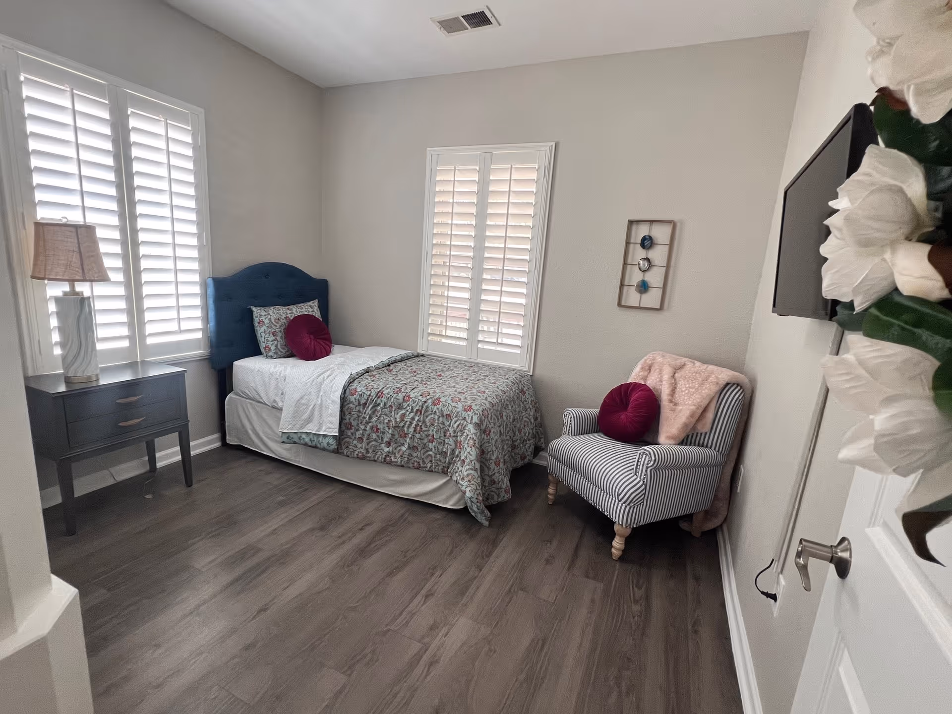 A small, cozy bedroom with a single bed featuring a blue upholstered headboard, floral bedding, and decorative pillows. Next to the bed is a dark wooden nightstand with a lamp. A striped armchair with a pink throw blanket and a round pink pillow is placed near the wall. The room has two windows with white plantation shutters and wood flooring.