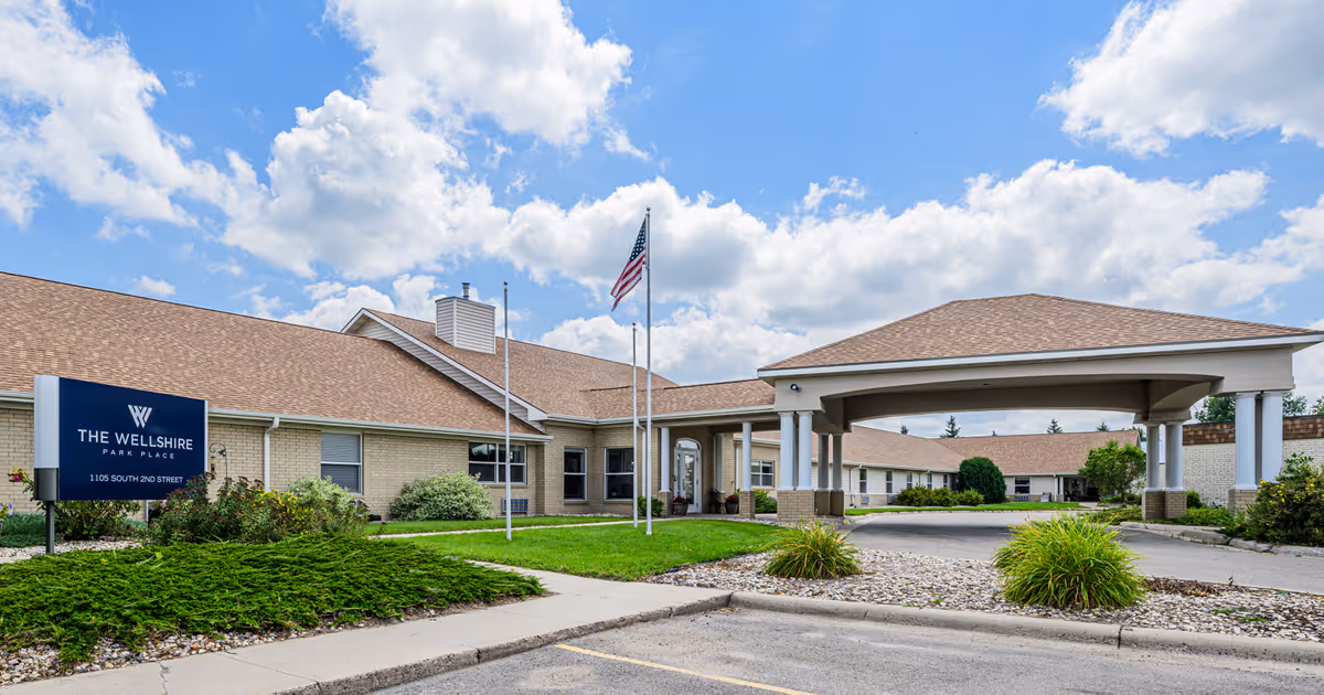 Exterior front entrance of The Wellshire Park Place senior living facility with a covered porte-cochere, flagpoles, sign, and landscaped grounds under a blue sky.