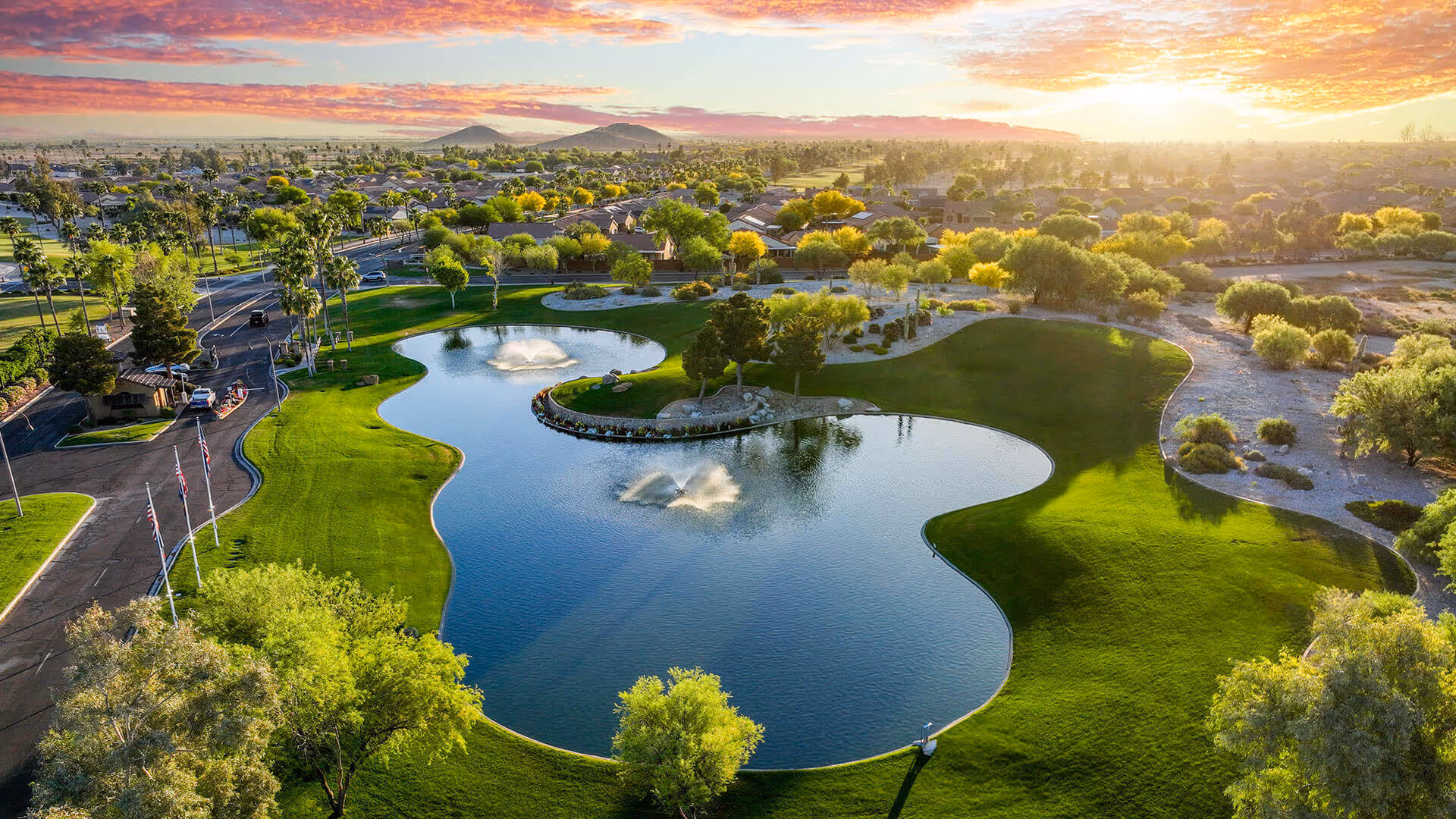 Aerial view of a landscaped pond with fountains, manicured lawns and trees in a residential community at sunset.