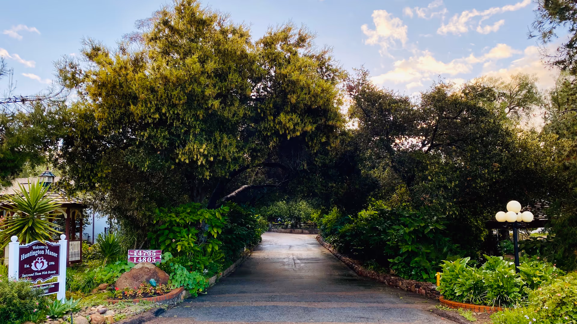 Entrance driveway to Huntington Manor facility surrounded by lush green trees and plants, with a sign on the left reading 'Welcome to Huntington Manor Est. 1996' and a street address sign nearby, under a partly cloudy sky.