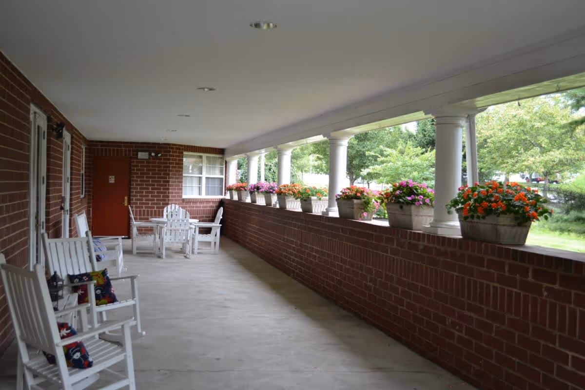 Covered outdoor patio area with white rocking chairs and a table with chairs. The patio has a brick half-wall with flower pots containing colorful flowers on top. White columns support the roof, and there is greenery visible beyond the patio.