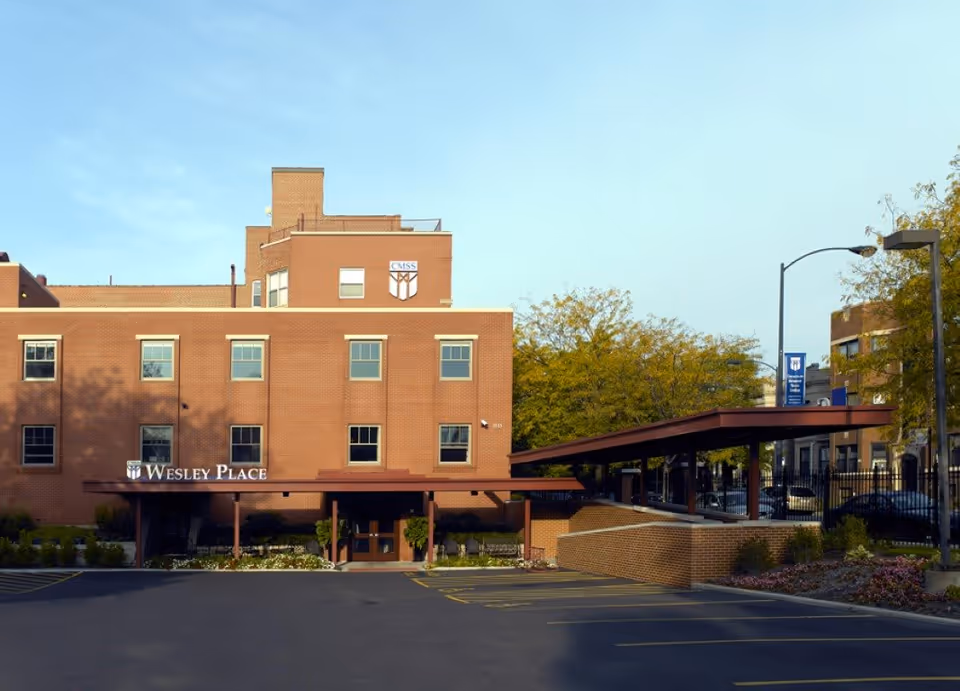 Exterior view of a brick building named Wesley Place with a covered entrance and a parking lot in front. Trees with autumn foliage and a street lamp are visible nearby under a clear sky.