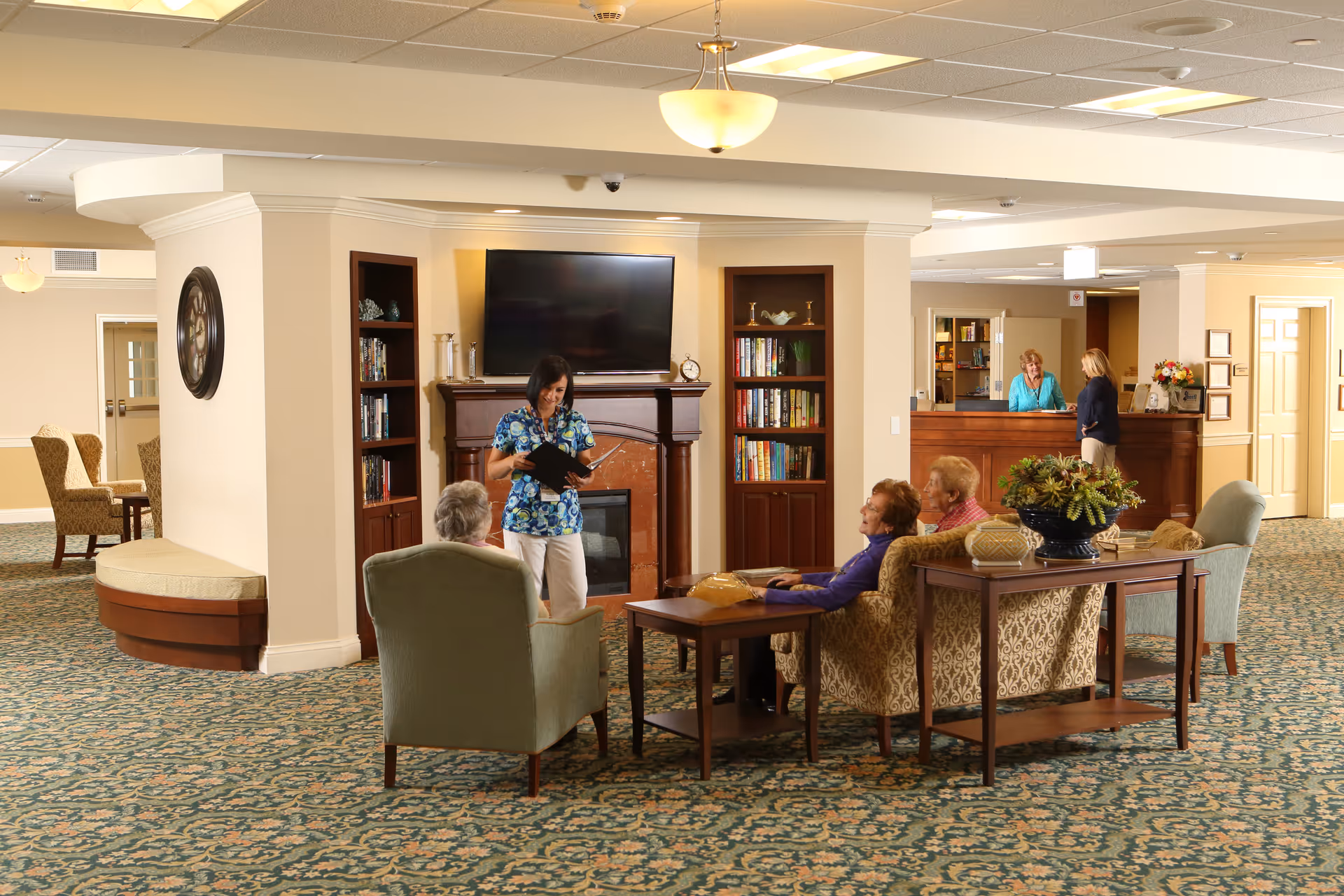 A cozy senior living facility common area with a group of elderly people seated on armchairs and sofas around a coffee table. A staff member is standing and holding a book or folder, engaging with the seated individuals. The room features a fireplace with a TV mounted above it, built-in bookshelves, and a patterned carpet. In the background, two women are conversing at a reception desk.