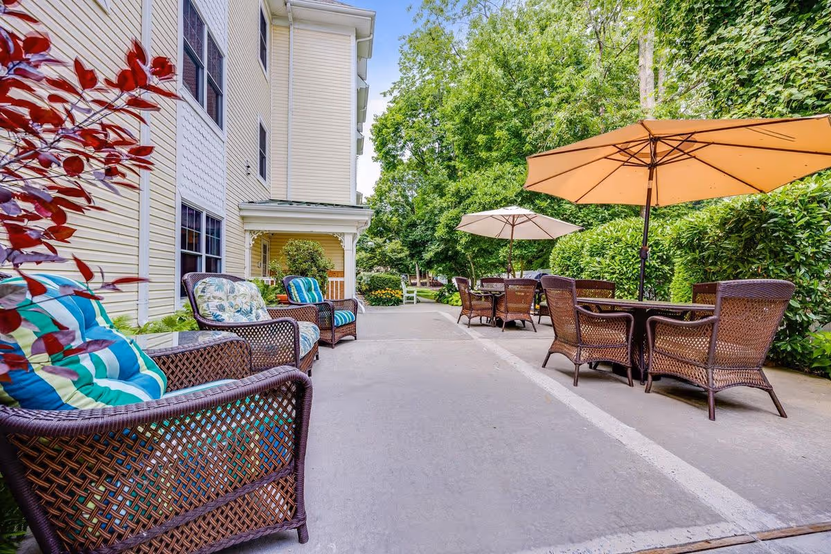 Outdoor patio area at Sunrise of Glen Cove with wicker chairs featuring colorful cushions, tables with large beige umbrellas, surrounded by greenery and trees next to a light yellow building.