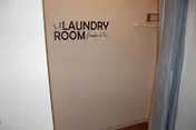 A small laundry room with a white wall featuring the text 'The Laundry Room loads of fun' in black letters. There is a shelf with a basket on the right side and a curtain partially visible on the right edge.
