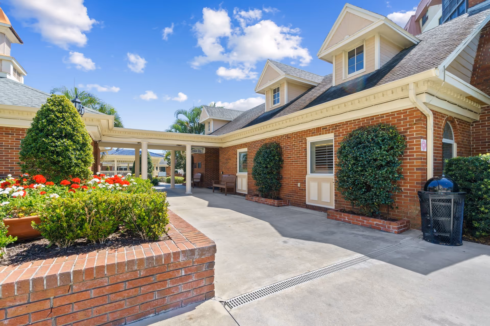 Exterior view of a senior living facility with brick buildings, manicured bushes, and flower beds under a blue sky with scattered clouds. There is a covered walkway with columns and benches along the building walls.