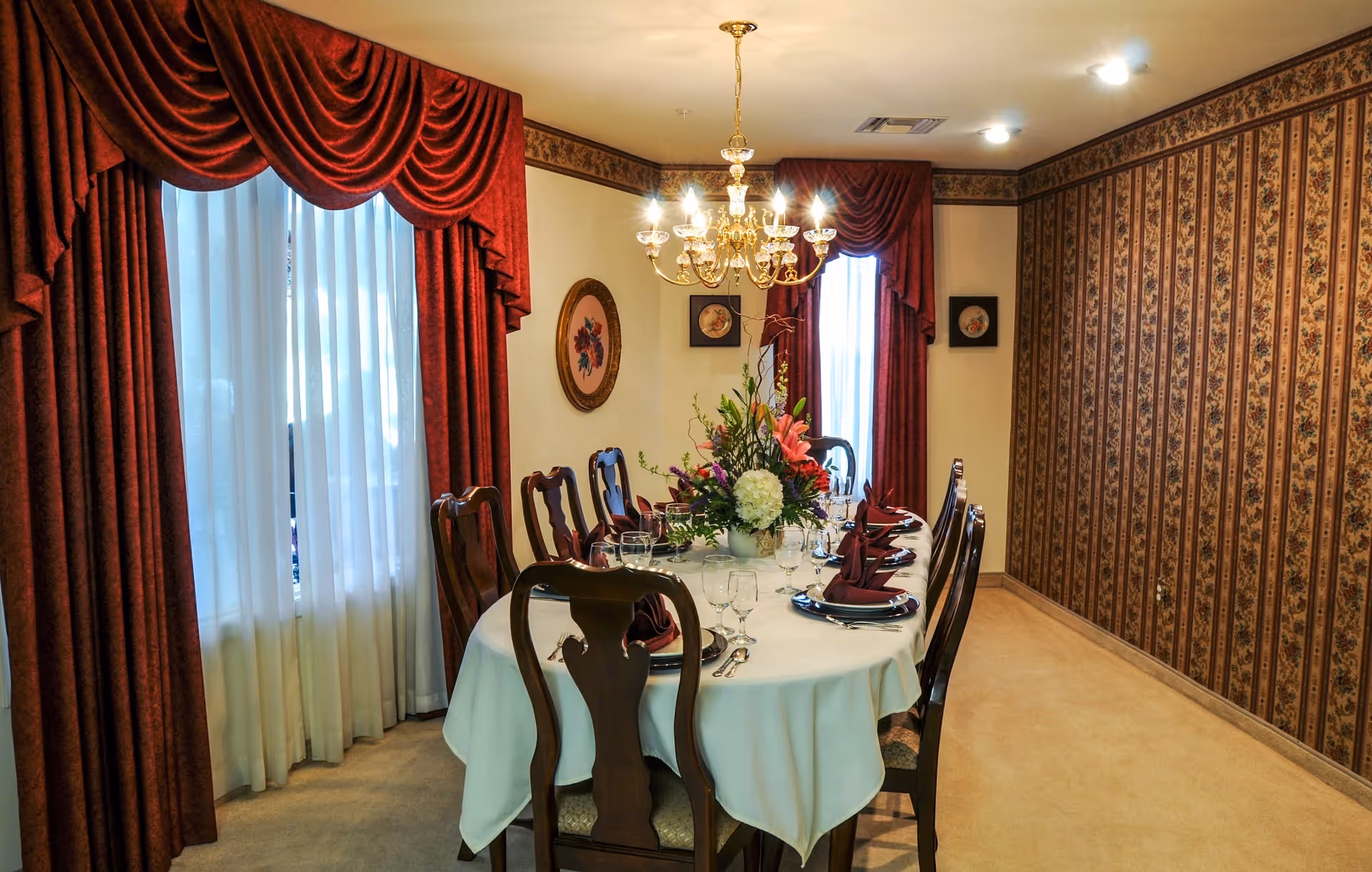 A formal dining room with a long oval table covered with a white tablecloth, set with plates, wine glasses, and folded burgundy napkins. The room features ornate wooden chairs, red draped curtains over windows, floral wallpaper on one wall, and a chandelier hanging from the ceiling. A floral centerpiece decorates the middle of the table.