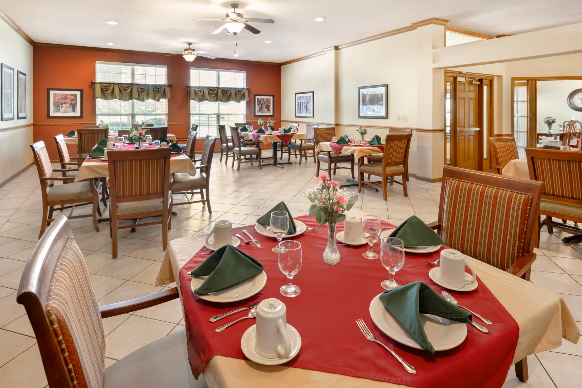A bright dining room with multiple tables set for a meal. Each table has a red tablecloth with beige underlay, green folded napkins, white plates, cups, and glassware. There are wooden chairs with striped upholstery around the tables. The room has large windows with green valances, framed artwork on the walls, and ceiling fans with lights.