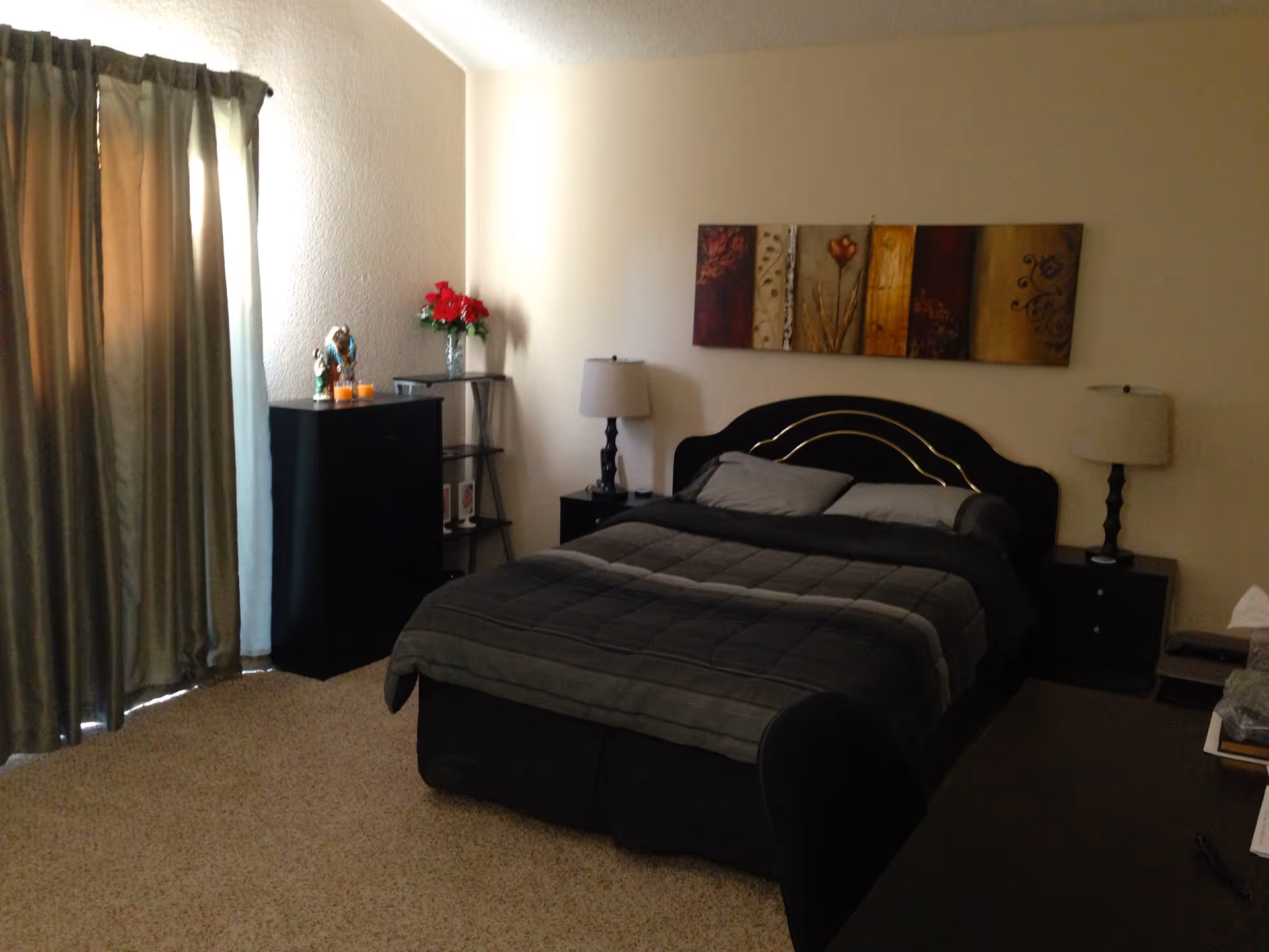 A bedroom with a double bed covered with a dark gray comforter and two pillows. On either side of the bed are black nightstands with matching lamps. Above the bed hangs a multi-panel decorative wall art featuring various colors and floral designs. To the left, there is a black dresser with decorative items and a vase of red flowers on top. Beige curtains cover a window or sliding door, and the floor is carpeted in a light beige color.