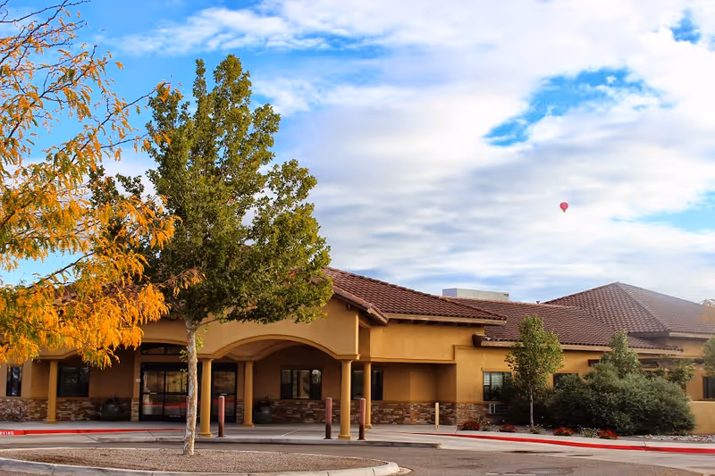 Exterior view of a single-story building with a tiled roof and an arched entrance, surrounded by trees with green and autumn-colored leaves under a partly cloudy sky with a hot air balloon in the distance.