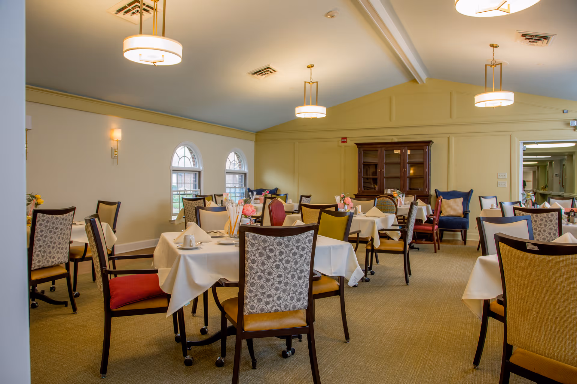 A dining room in a senior living facility with multiple tables covered in white tablecloths, set with napkins, cups, and small flower arrangements. The room has a beige carpet, light yellow walls, arched windows, and several chairs with patterned upholstery. There are three ceiling lights and a wooden cabinet against the far wall.