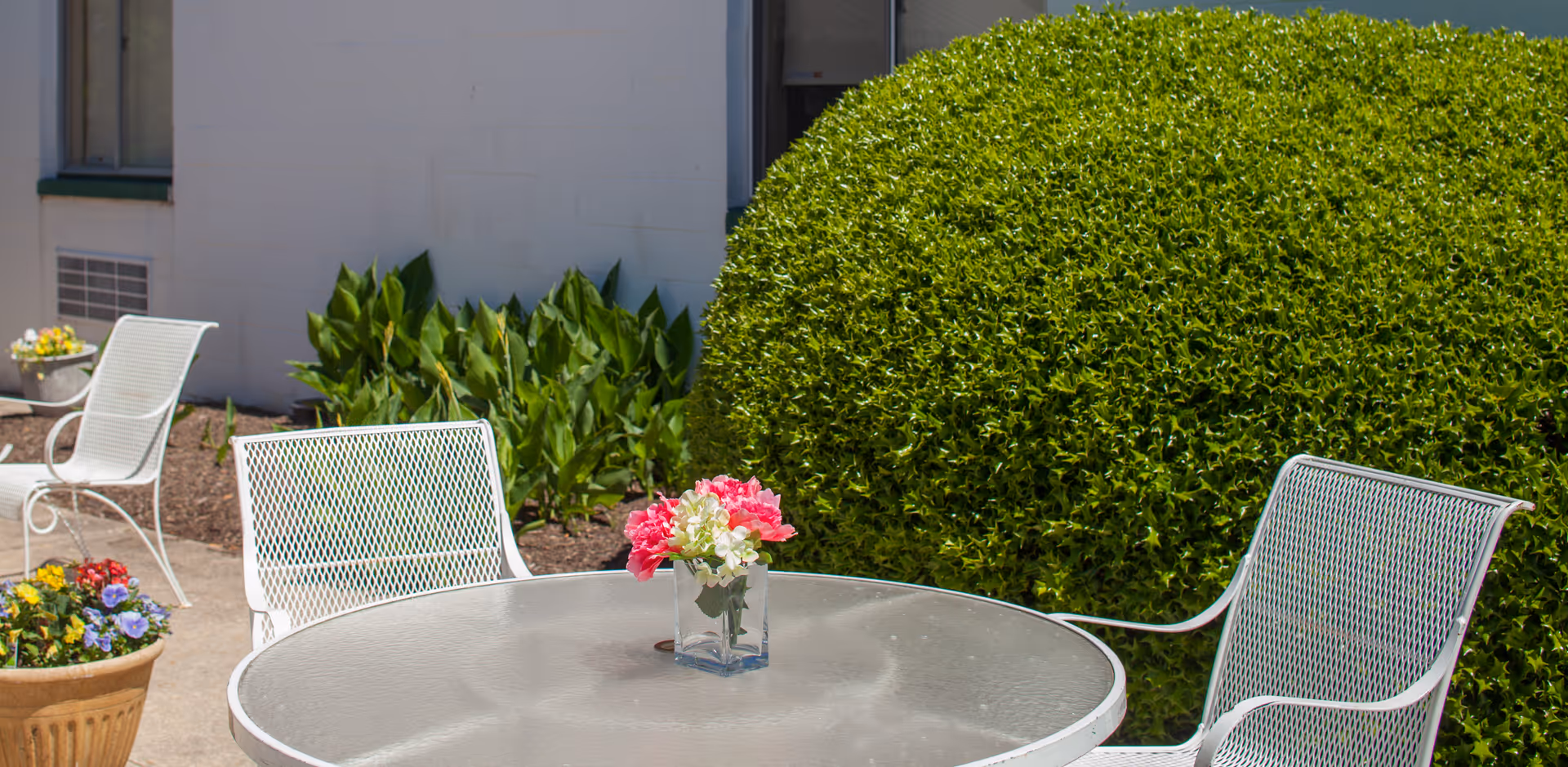 Outdoor patio area with a round glass table and white metal chairs. A small vase with pink and white flowers is placed on the table. There are green bushes and plants in the background along with a building wall.