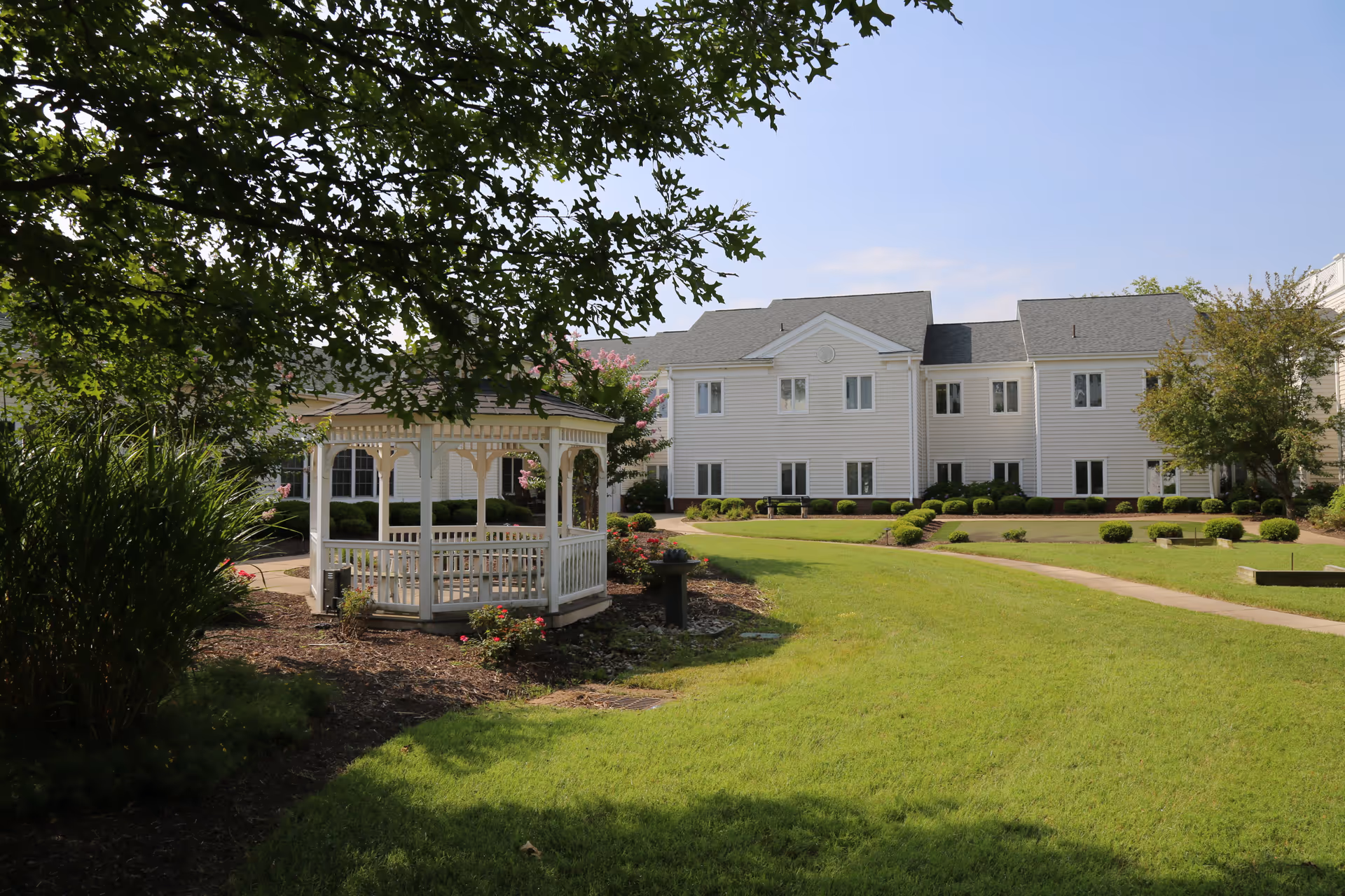 A well-maintained outdoor garden area at Lucy Corr featuring a white wooden gazebo surrounded by green grass, bushes, and trees with a large white building in the background under a clear blue sky.