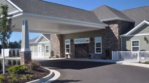 Entrance of a retirement living facility with a covered driveway, stone and green siding exterior walls, white railings, and landscaped garden beds under a clear blue sky.