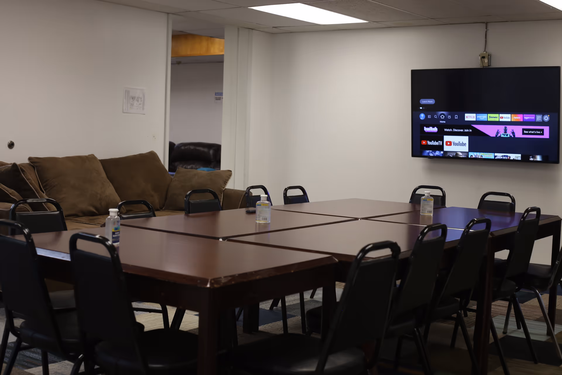A meeting/activity room with several tables pushed together surrounded by chairs, a wall-mounted TV, and a couch in the background.