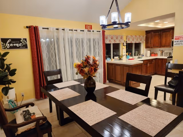 Dining room with a dark wooden table set with placemats and a floral centerpiece, yellow walls, curtains, and an adjoining kitchen.