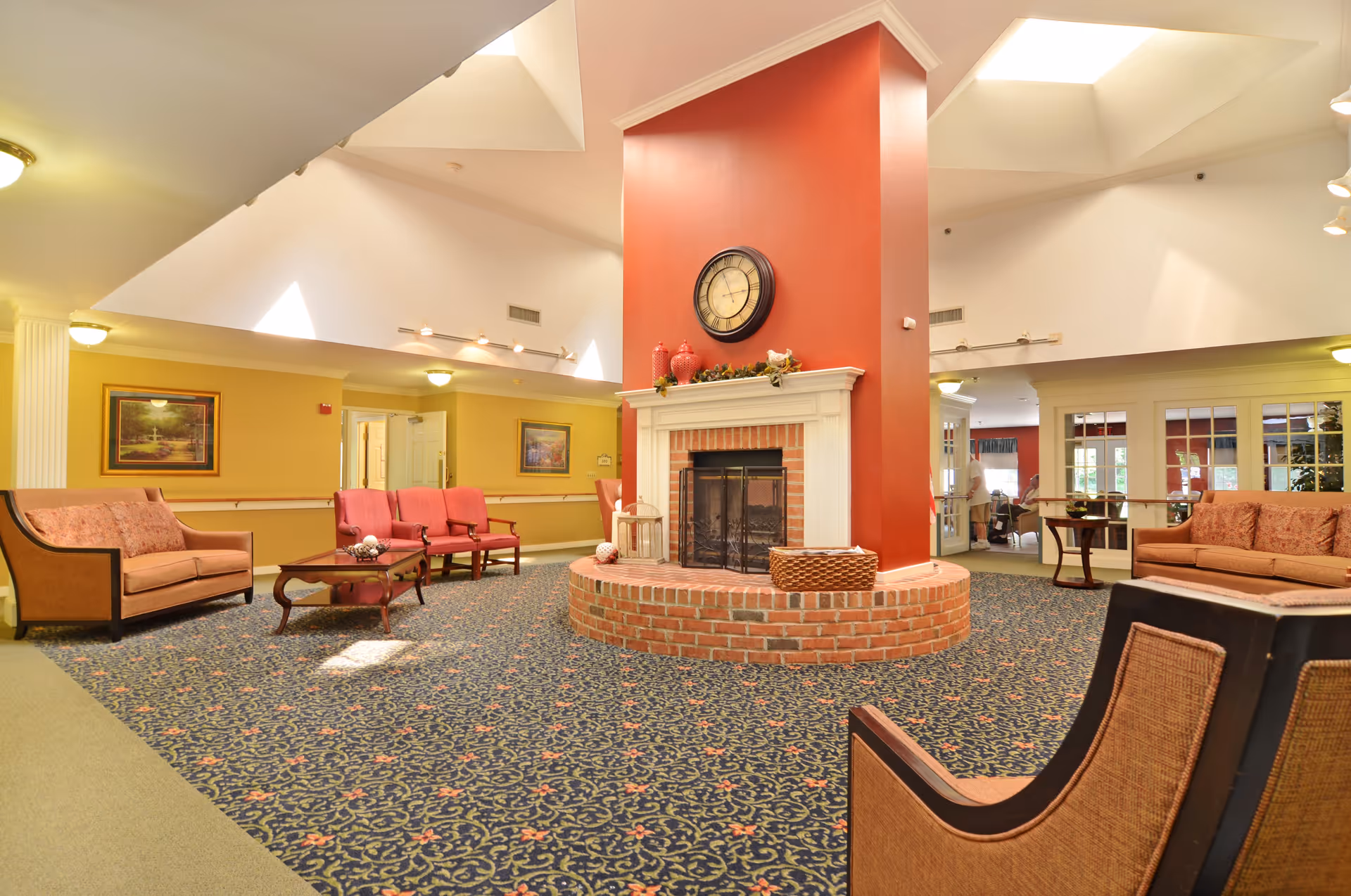 A spacious senior living facility common area with a central brick fireplace featuring a red accent wall and a large clock above it. The room is furnished with multiple upholstered chairs and sofas in shades of pink and beige, arranged around coffee tables on a patterned carpet. The walls are painted yellow with framed artwork, and the ceiling has skylights allowing natural light to brighten the space.