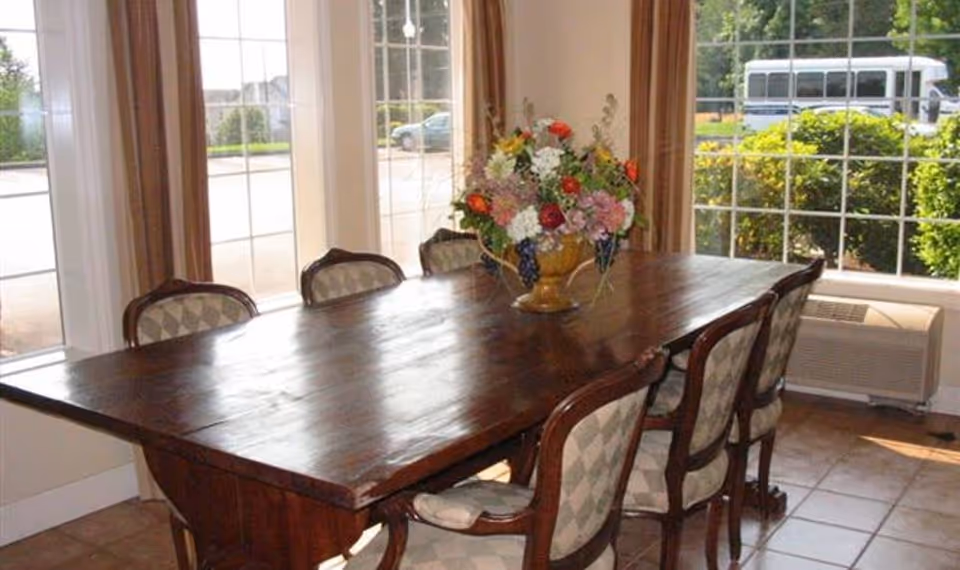 A wooden dining table with six upholstered chairs arranged around it, placed in a room with large windows showing greenery and a parked vehicle outside. A decorative floral arrangement is centered on the table.