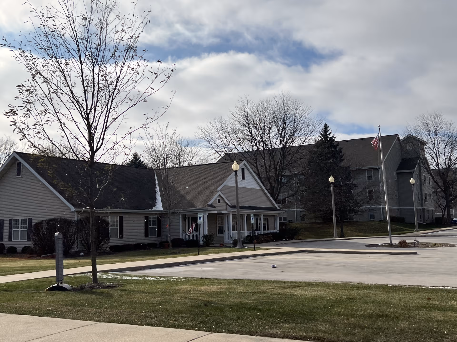 Exterior view of a senior living facility with a single-story building in the foreground and a larger multi-story building in the background. There are leafless trees, a flagpole with an American flag, street lamps, and a paved parking area surrounded by grass.
