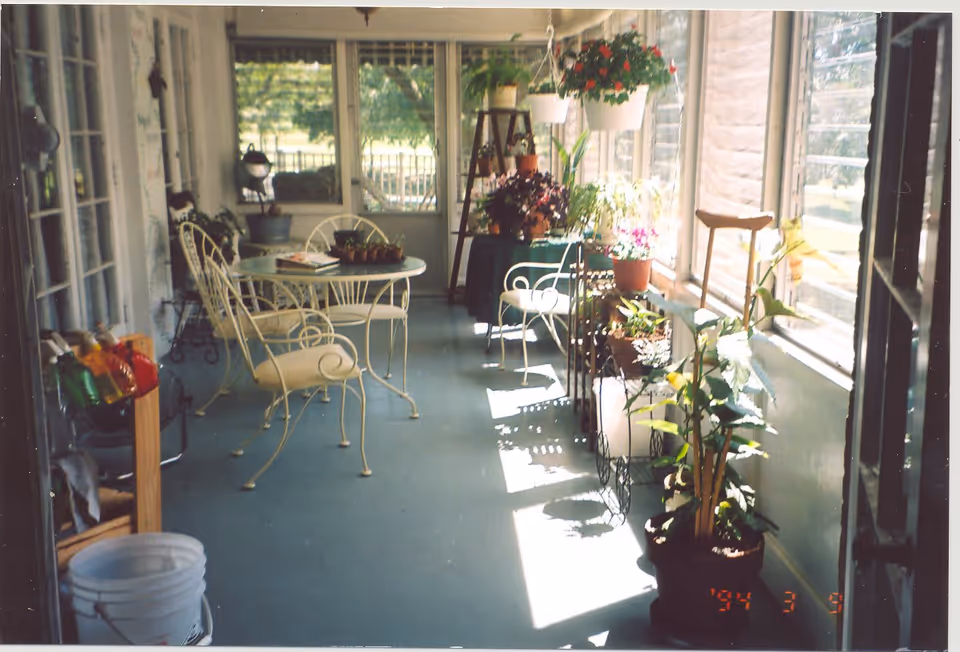 Sunroom with wrought-iron table and chairs and numerous potted plants along large windows.