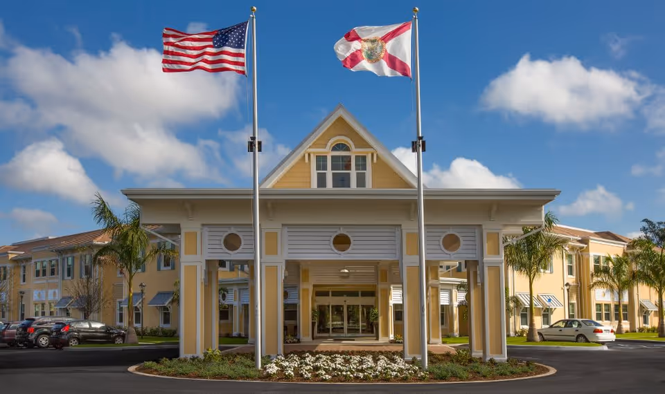 Front exterior view of a senior living facility building with two flagpoles displaying the American flag and the Florida state flag. The building is painted yellow with white trim and has a covered entrance with circular windows. There are palm trees and parked cars visible on either side of the entrance under a partly cloudy blue sky.