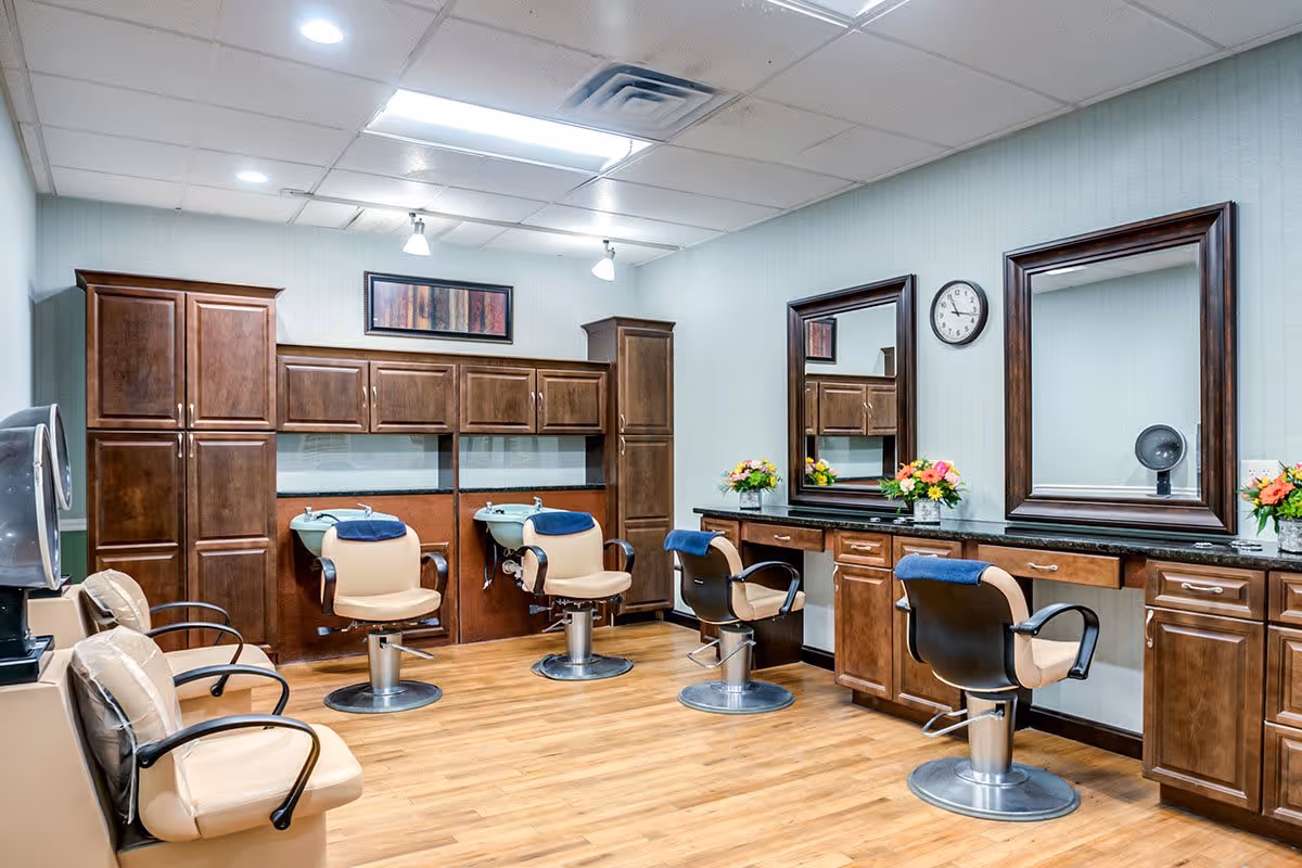 Interior view of a salon area with wooden cabinets, three salon chairs with blue towels on the headrests, two large mirrors on the wall, a clock, and flower arrangements on the counter. The floor is wooden and the ceiling has recessed lighting.