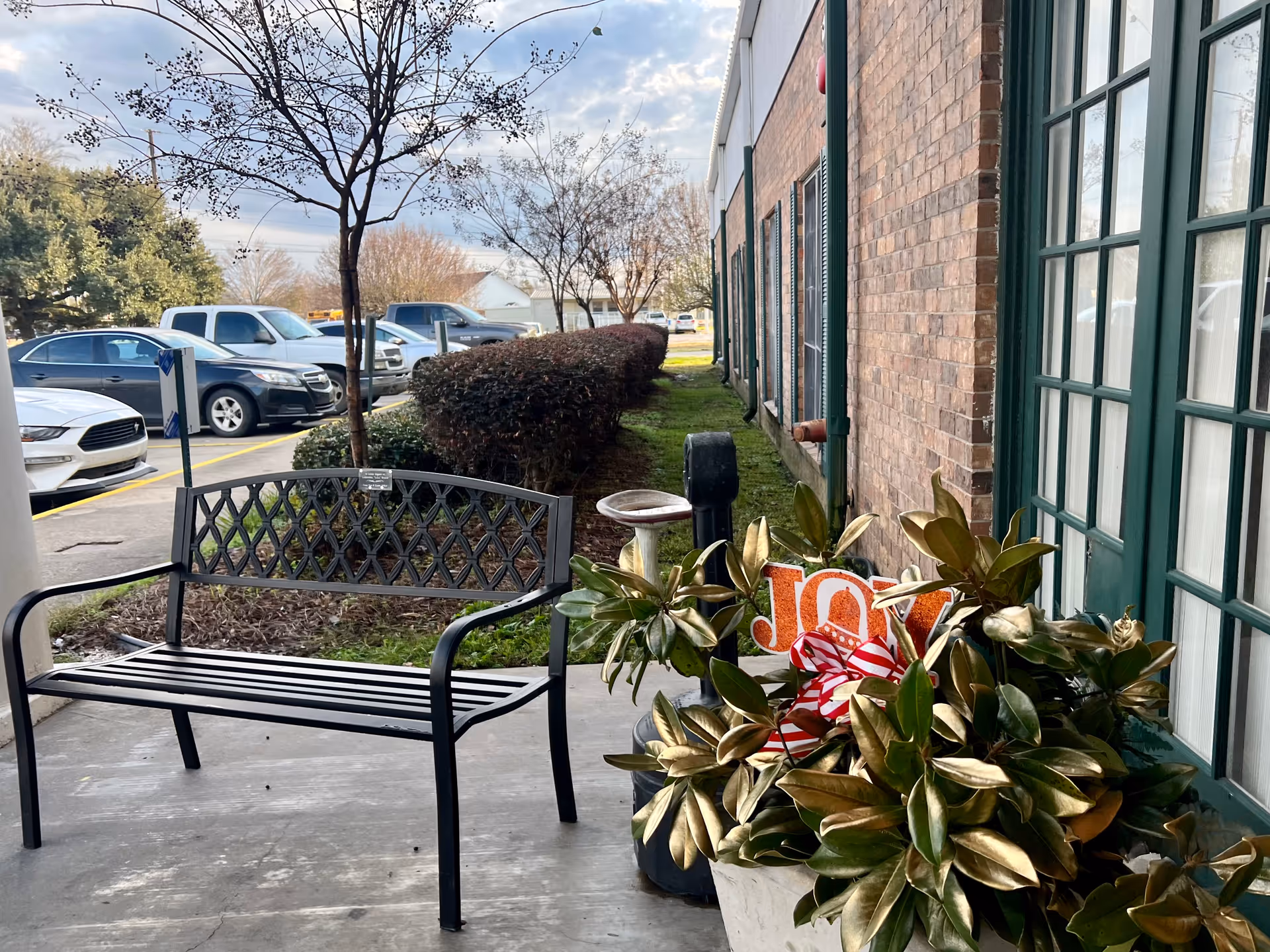 Outdoor seating area with a black metal bench on a concrete patio next to a brick building with green-framed windows. There is a large potted plant with a decorative sign that says 'JOY' and a red and white striped bow. In the background, there are parked cars, trees, and bushes under a partly cloudy sky.