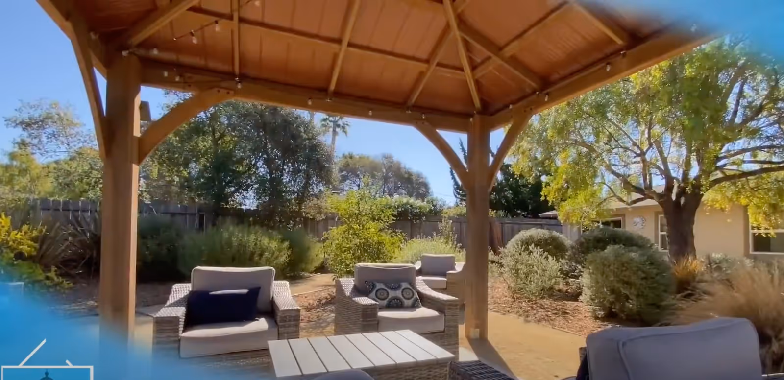 Outdoor seating area under a wooden gazebo with cushioned wicker chairs and a table, surrounded by trees, bushes, and a wooden fence in the background on a sunny day.