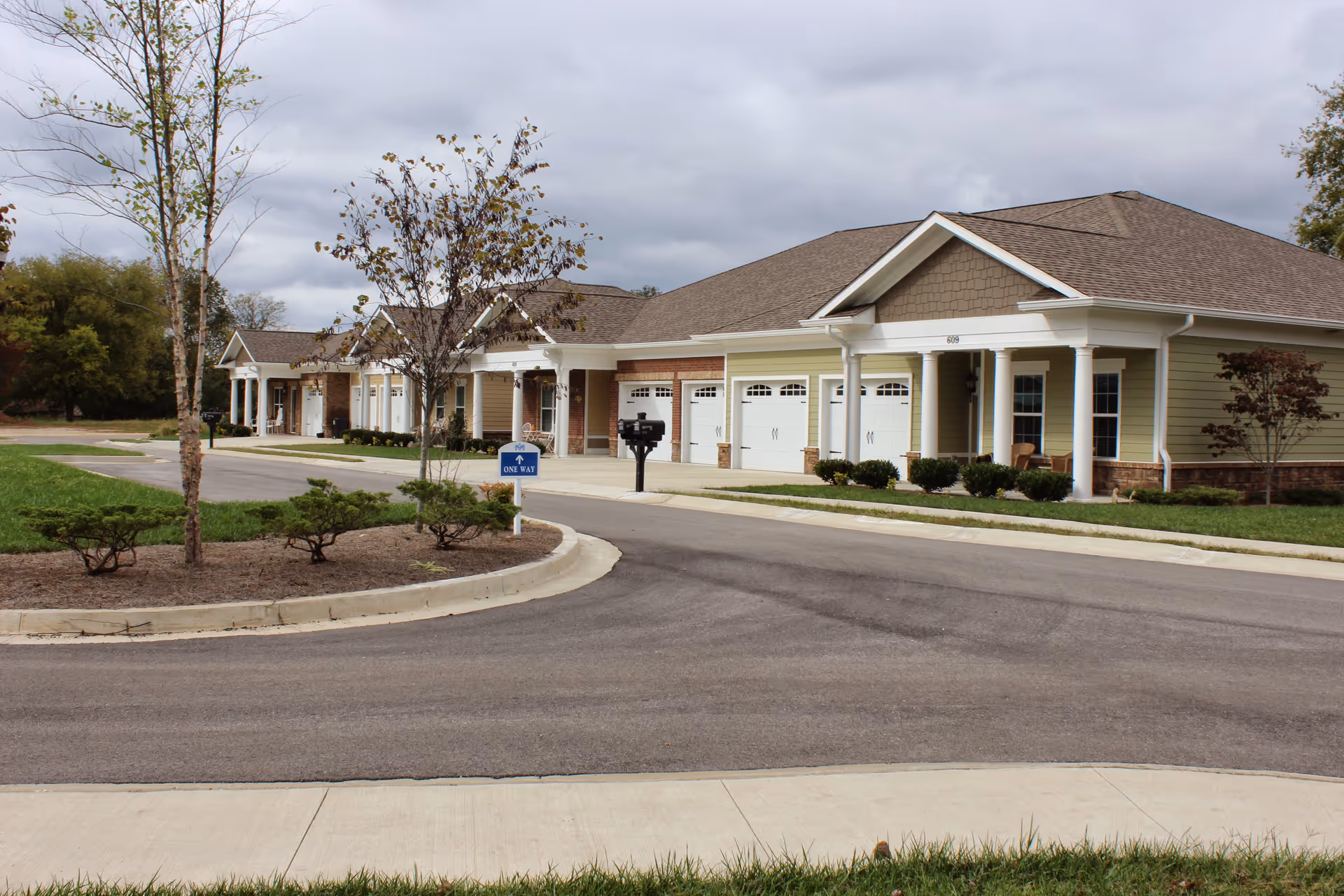 Exterior view of a single-story senior living facility building with multiple attached units, each having a garage door and a small porch area. The building is surrounded by a paved road, landscaped bushes, small trees, and a grassy area under a cloudy sky.