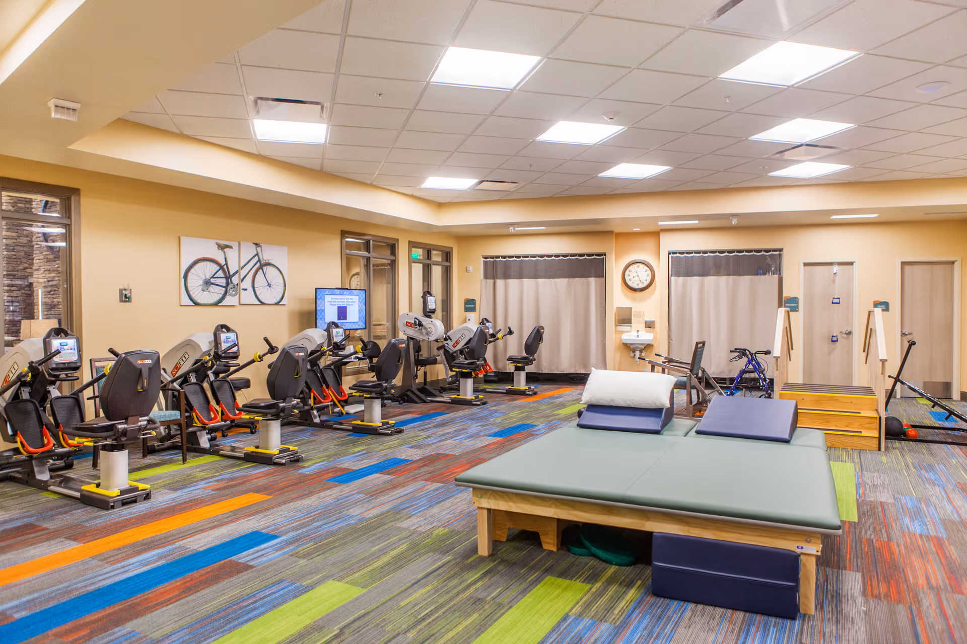 A brightly lit exercise room with several stationary recumbent bikes lined up against the left wall. In the center, there is a padded therapy table with cushions. The floor has a colorful patterned carpet with green, blue, orange, and gray stripes. On the back wall, there are two curtained areas, a clock, a sink, and some exercise equipment including a walker and a scooter. A picture of a bicycle is hung on the left wall above the exercise bikes.