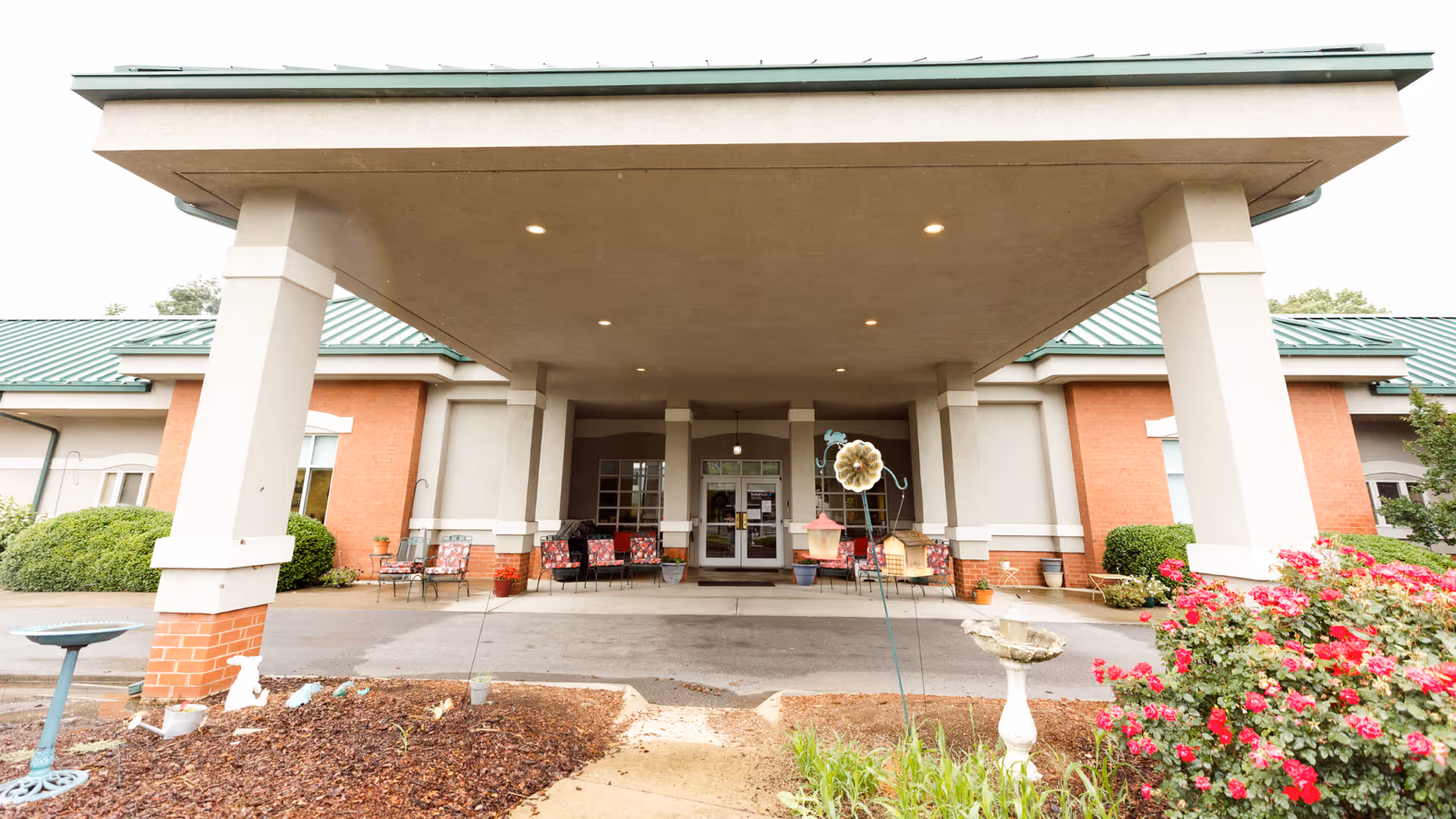 Front entrance of Madison House facility with a covered drop-off area supported by large columns. There are chairs with red cushions arranged near the entrance, potted plants, and a garden area with flowers and bird feeders in the foreground.