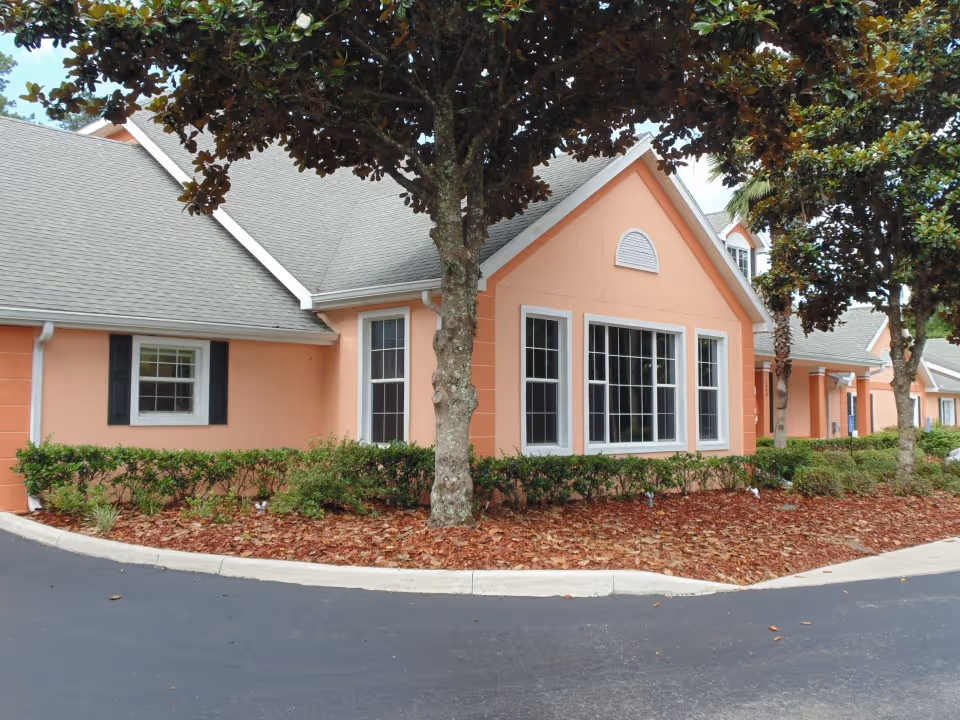 Exterior view of a peach-colored building with white trim and multiple windows, surrounded by landscaping with bushes and trees, and a paved driveway in front.