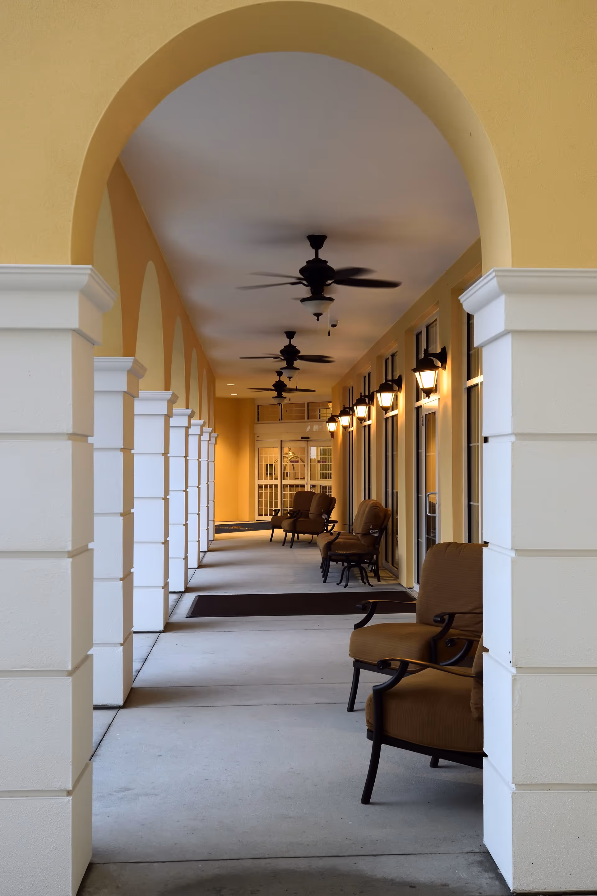Covered outdoor corridor with beige walls and white pillars, featuring several cushioned chairs along the right side, ceiling fans, and wall-mounted lantern-style lights leading to glass doors at the end.