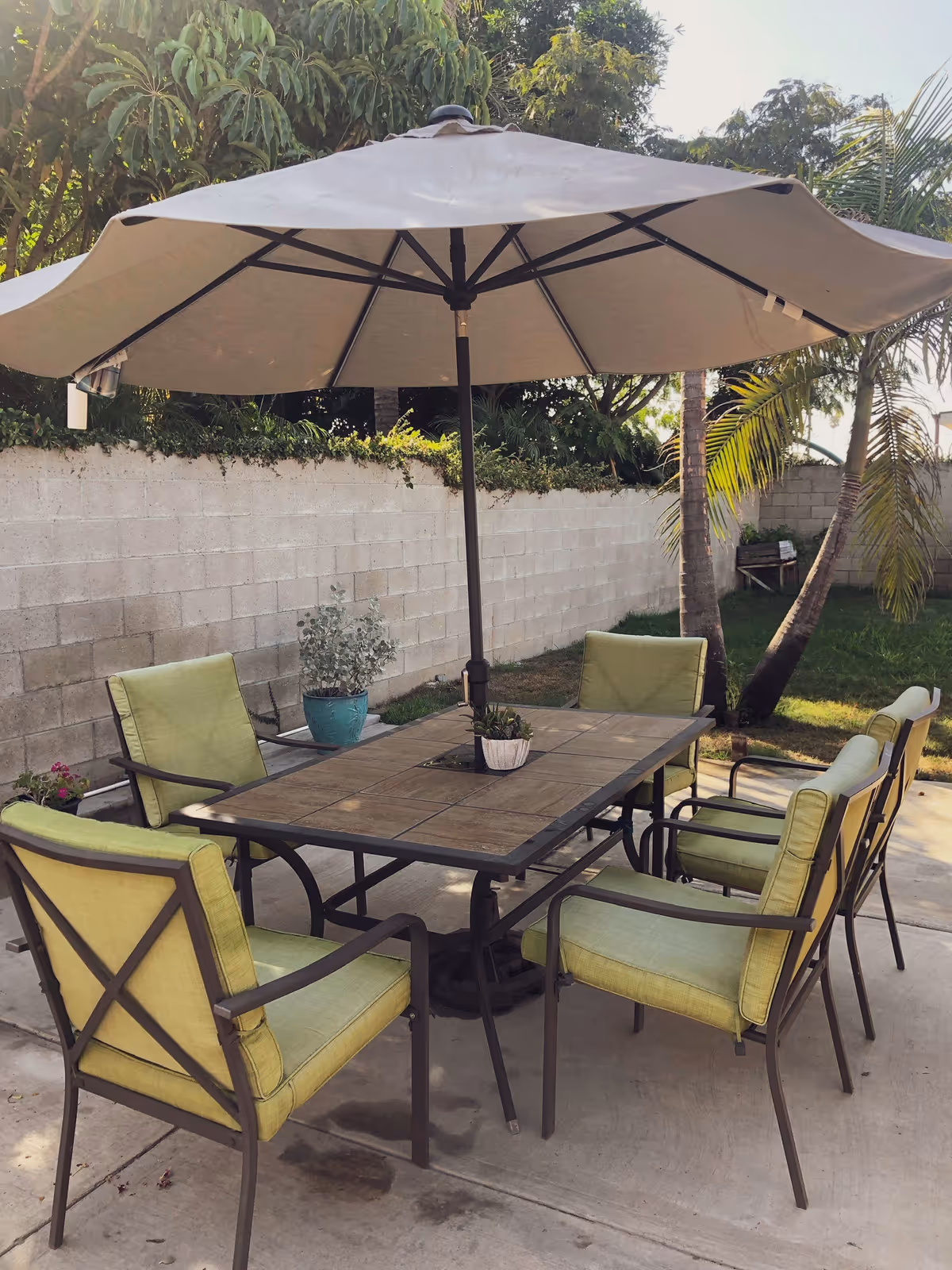 Outdoor patio area with a rectangular table and six green cushioned chairs arranged around it. A large beige umbrella is open above the table providing shade. The patio is surrounded by a concrete block wall, with trees and plants visible in the background.